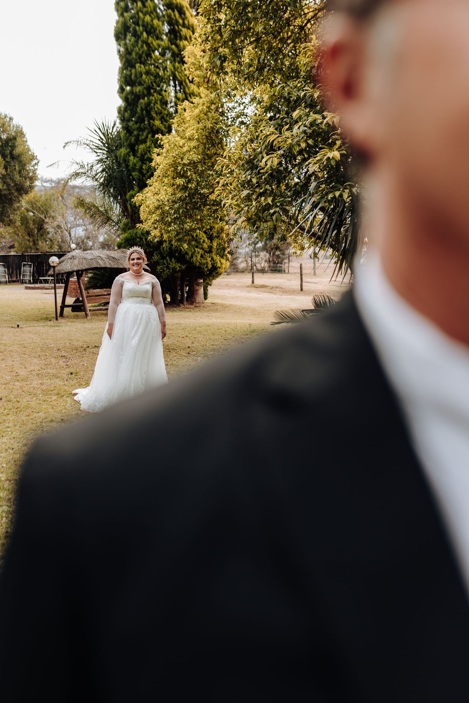 A bride in a white wedding gown stands in a garden setting while a suited figure is partially visible in the foreground.