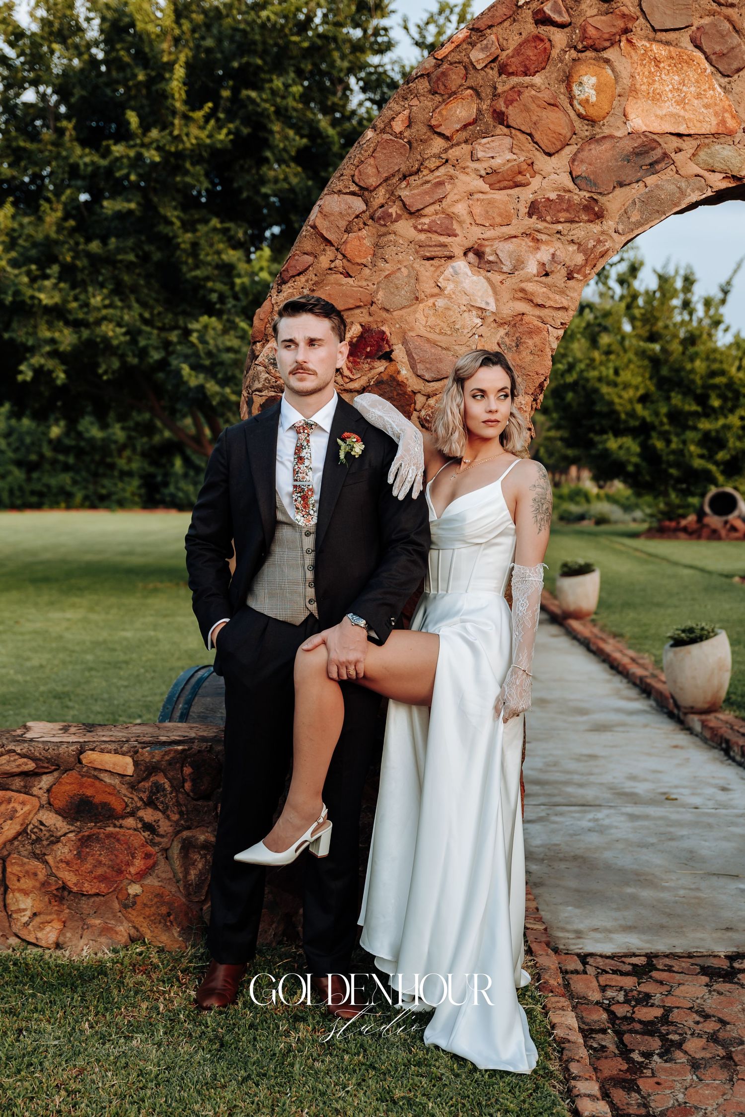 Wedding couple posing near rustic stone wall in an outdoor garden setting.