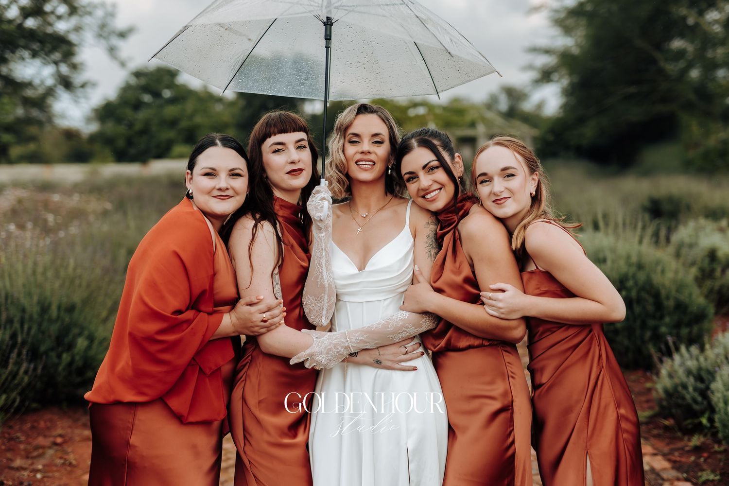 Wedding party poses with clear umbrella wearing white gown and rust colored bridesmaid dresses.