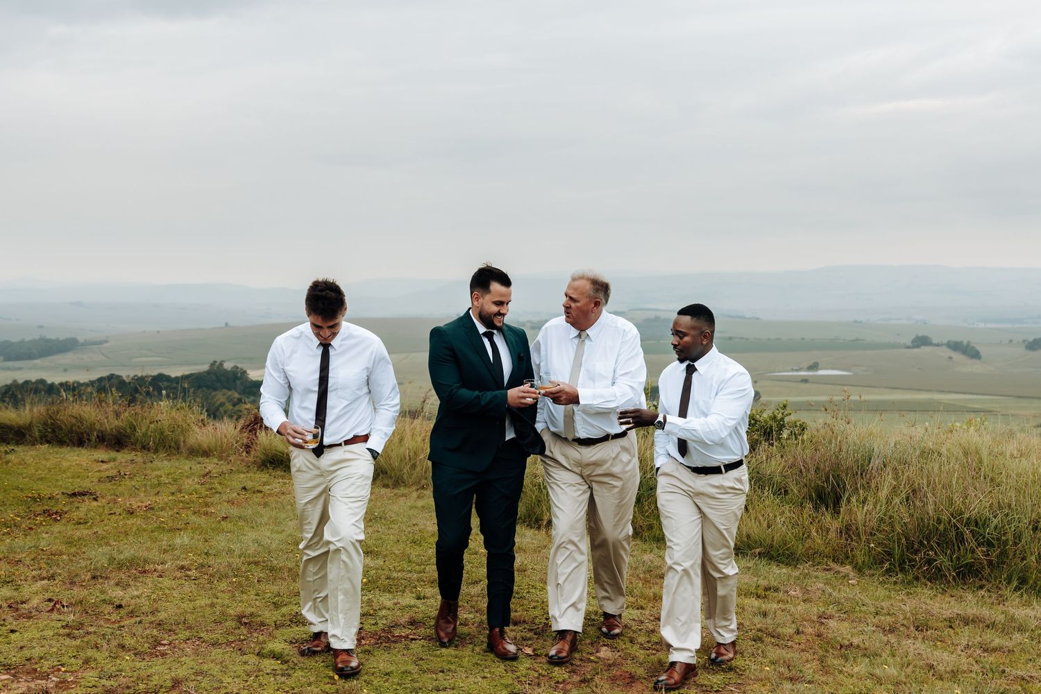 A group in formal attire stands together on a grassy hilltop overlooking a scenic valley landscape.