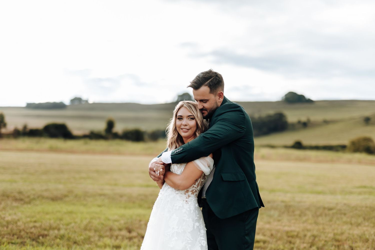 A couple in wedding attire embraces in a scenic field with rolling hills and golden grass in the background.