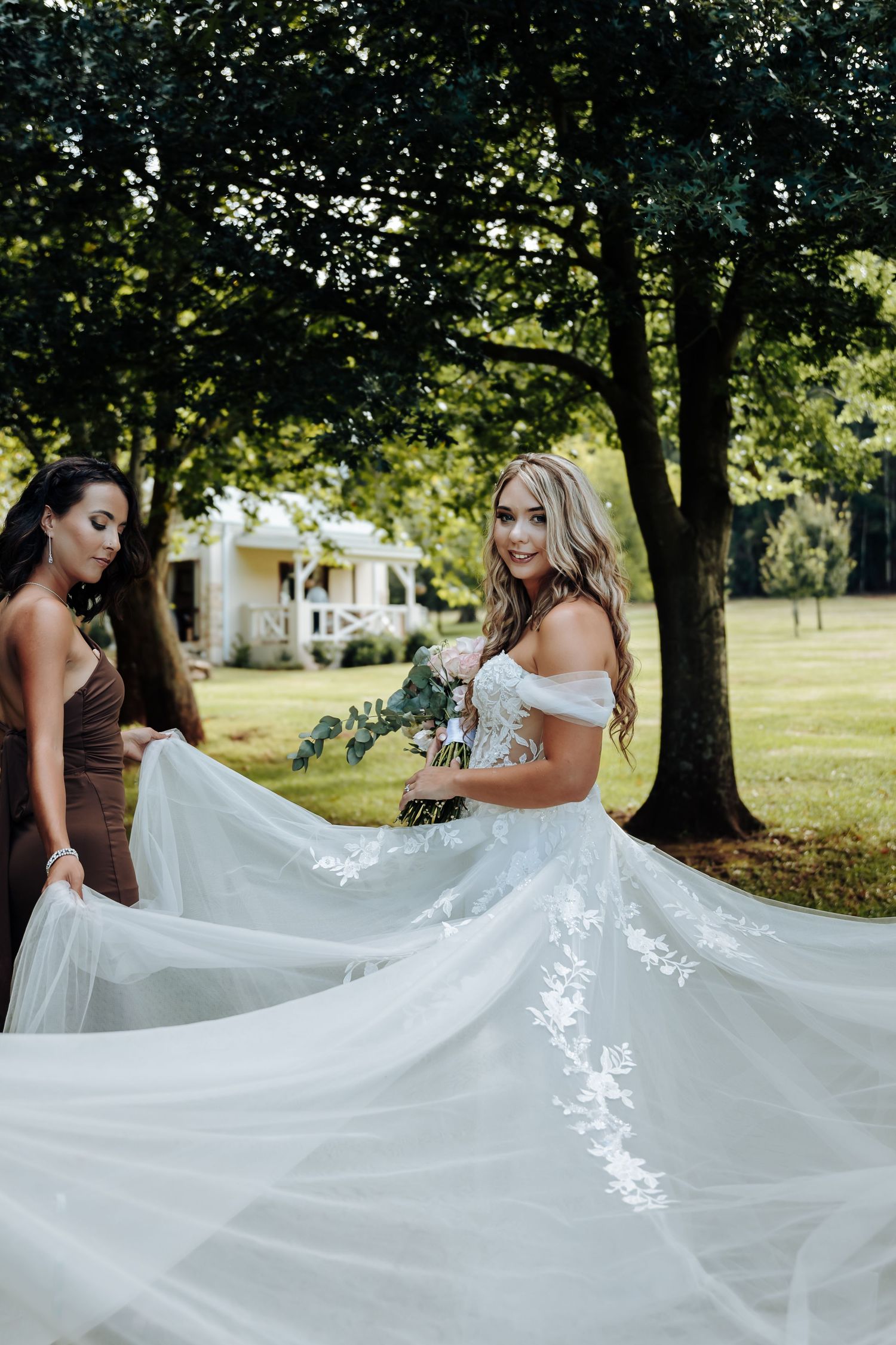 Bridesmaids in brown dresses help spread out a flowing white wedding dress train in an outdoor garden setting.