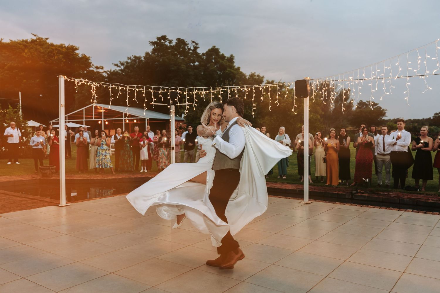 String lights illuminate an outdoor wedding reception venue at dusk with tables and chairs set up under a tent.