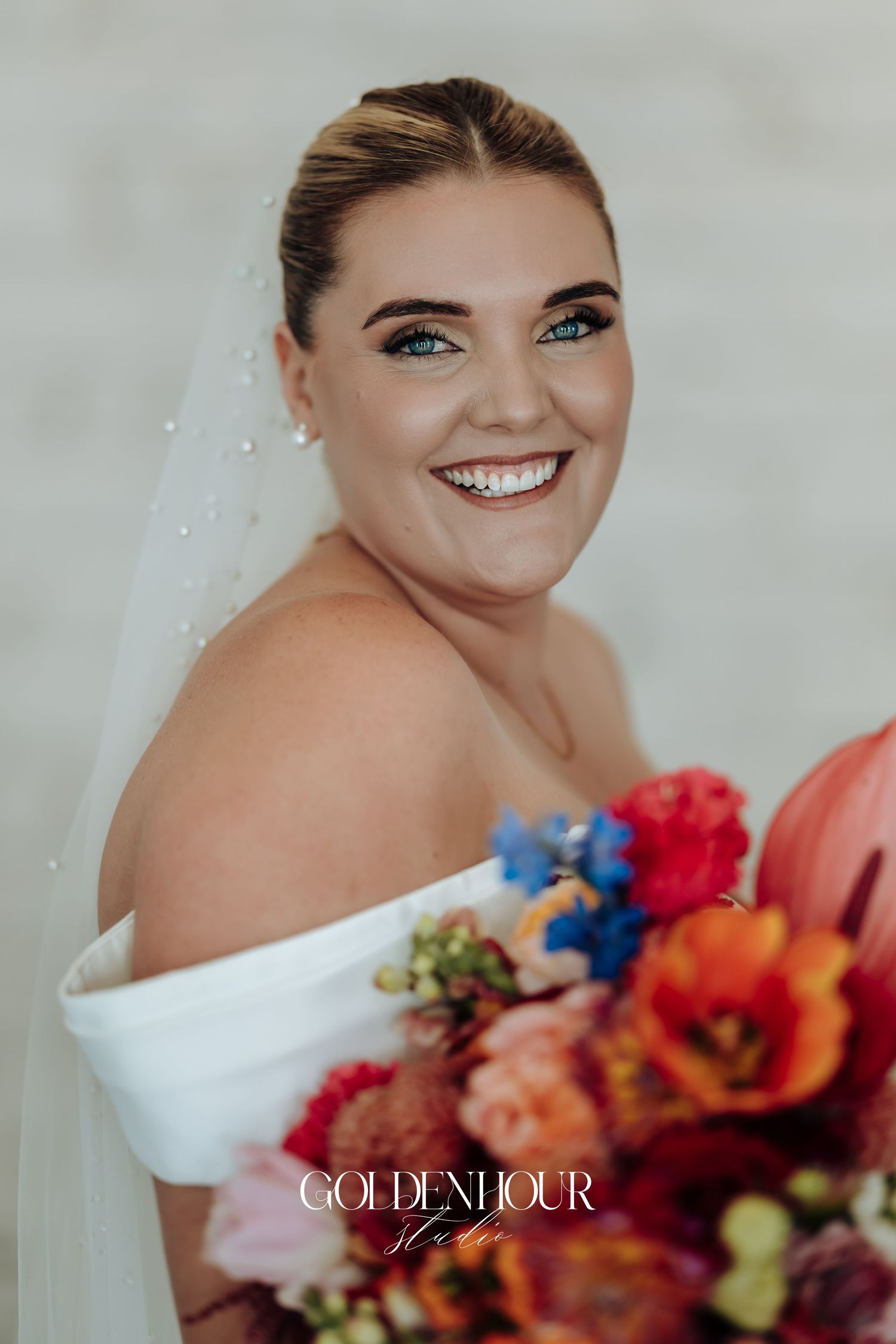 A bride in a white off-shoulder gown poses with vibrant red, blue and orange floral bouquet against a light backdrop.