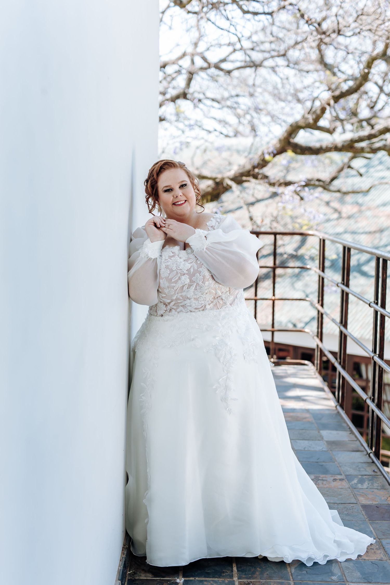 A bride in a white lace wedding gown poses outdoors under blooming cherry blossom trees on a spring day.