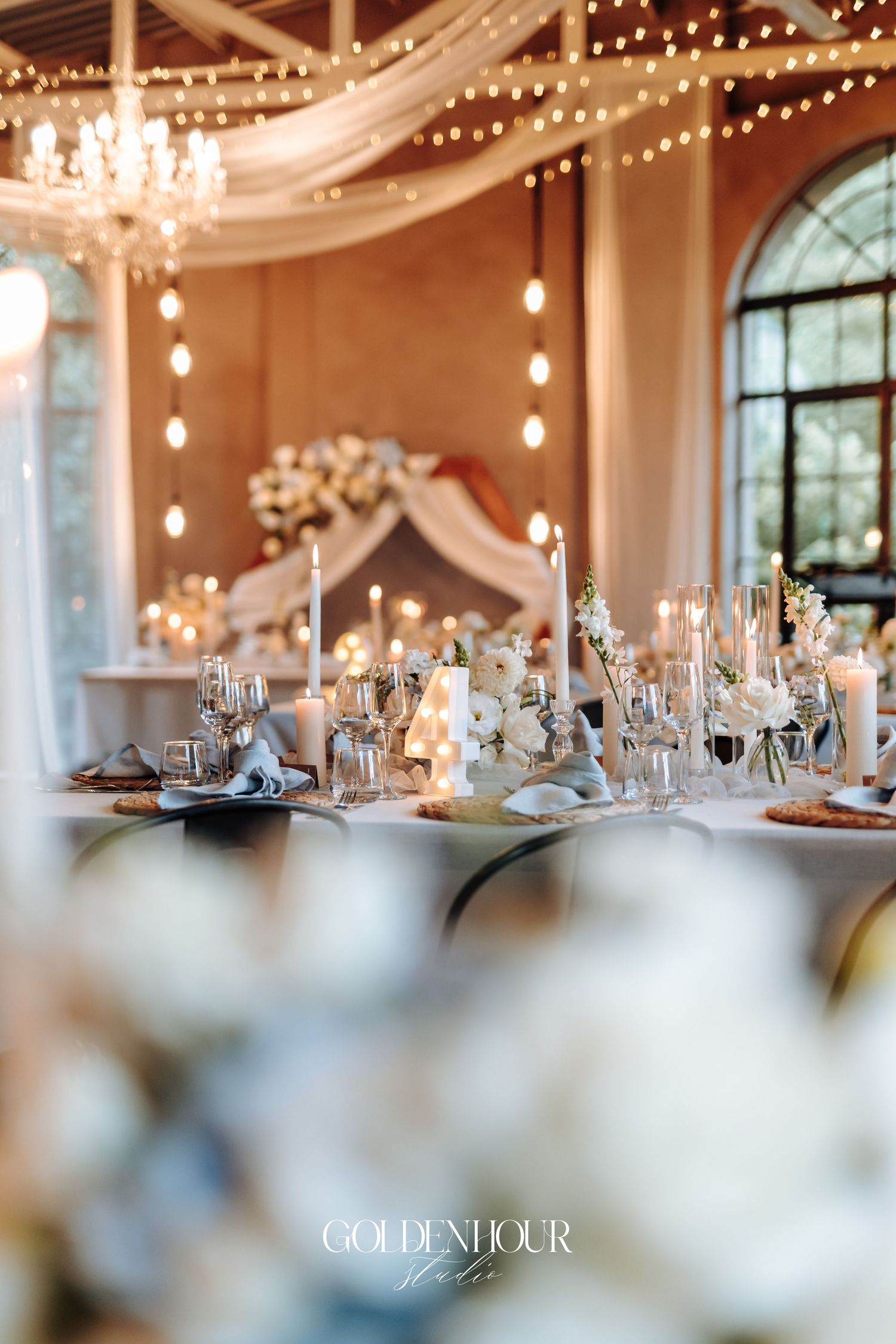 Elegant wedding reception tables set with glassware under twinkling string lights and white fabric draping.