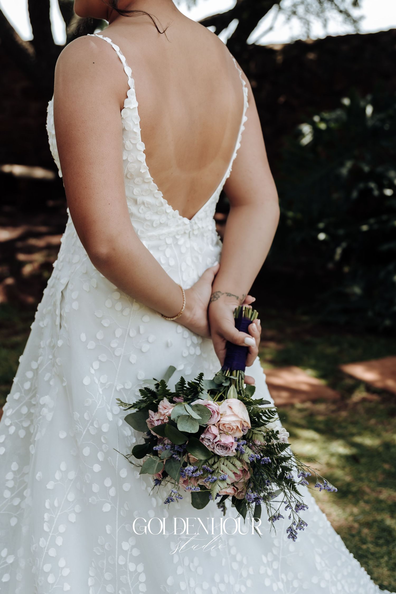 Back view of bride in white lace dress holding bouquet with pink roses and greenery.