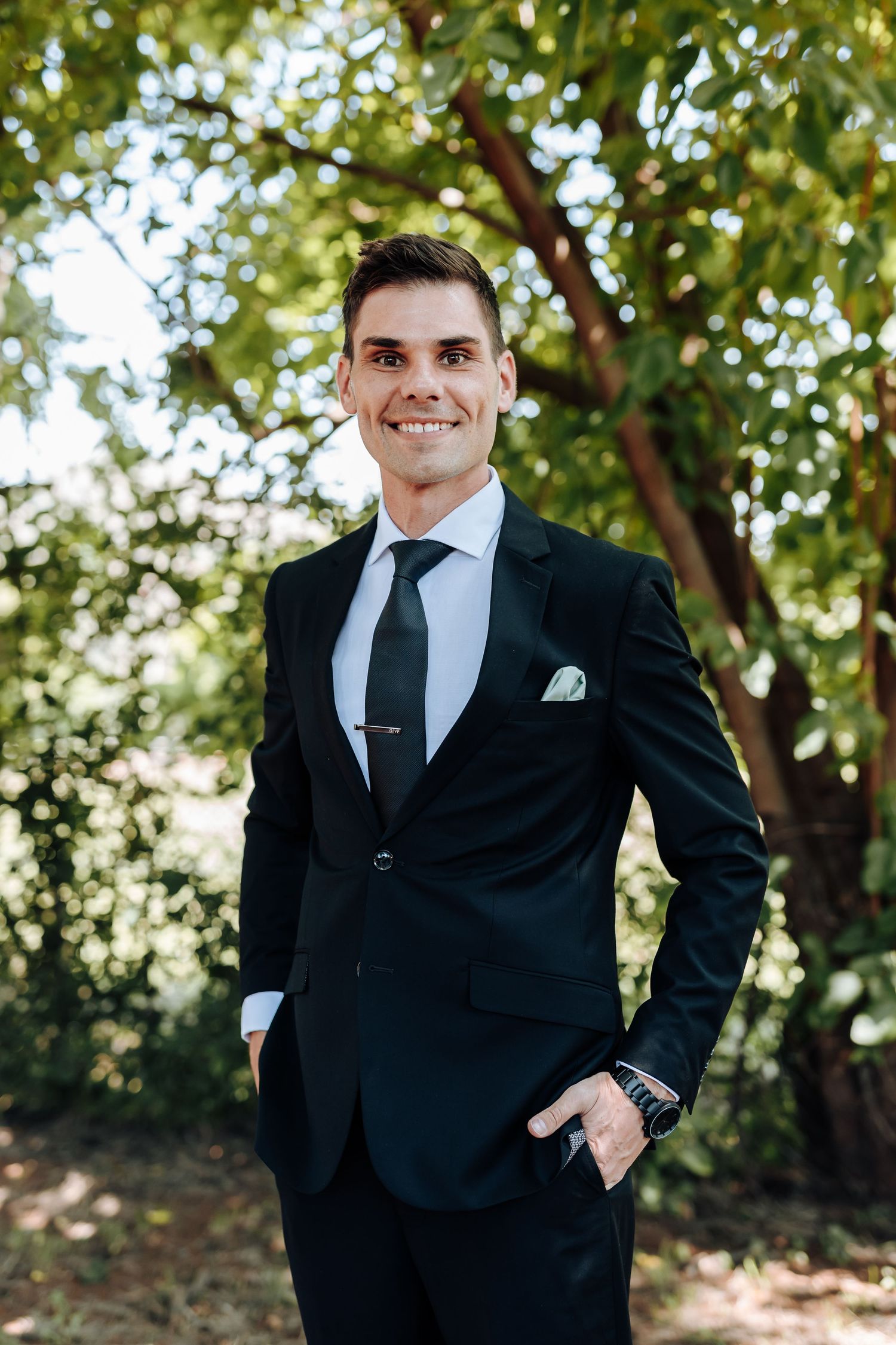 Wedding party member in black suit and tie posing against leafy background.