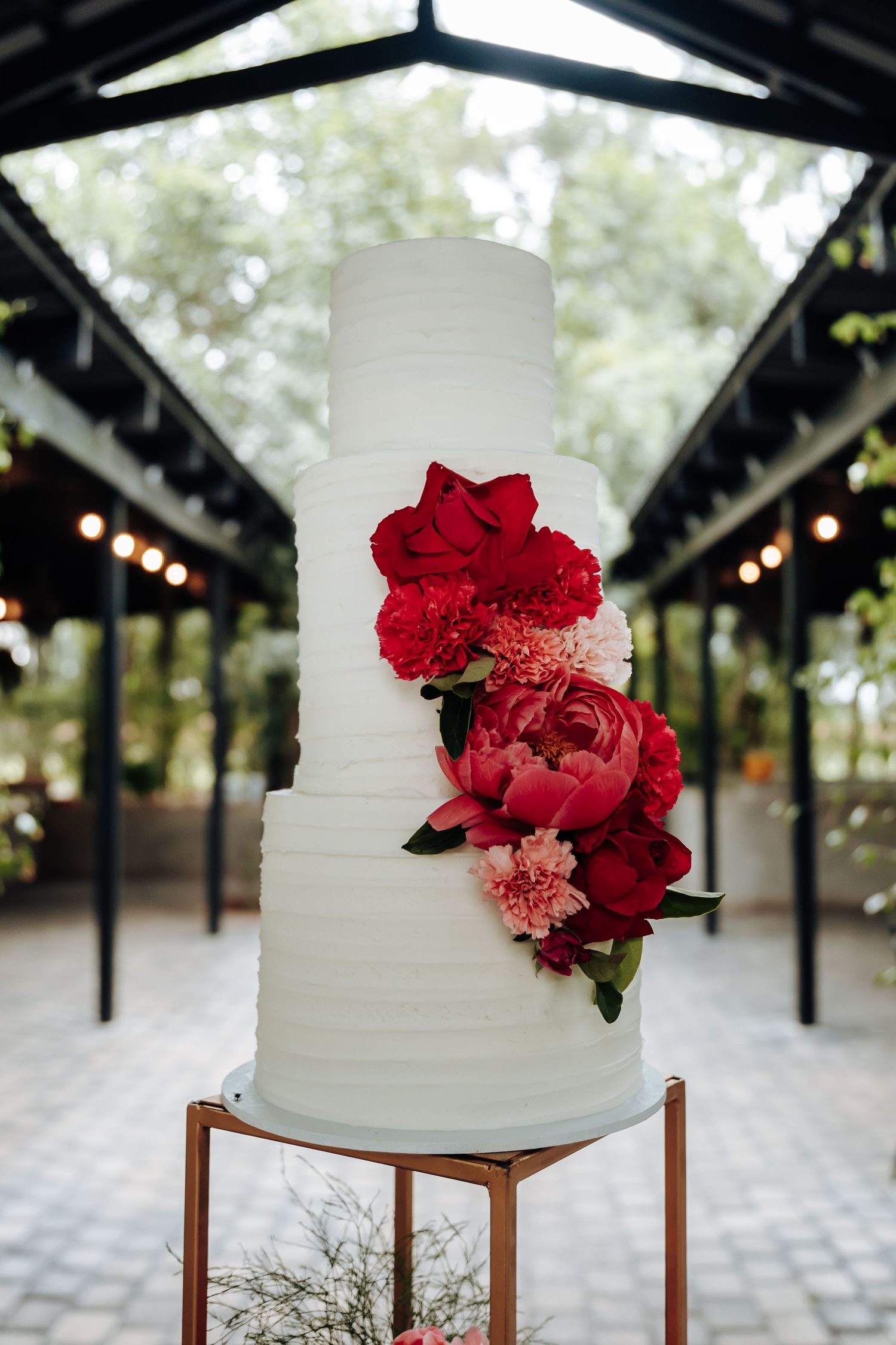 White wedding cake with cascading red roses displayed on wooden stand under rustic wooden pavilion.