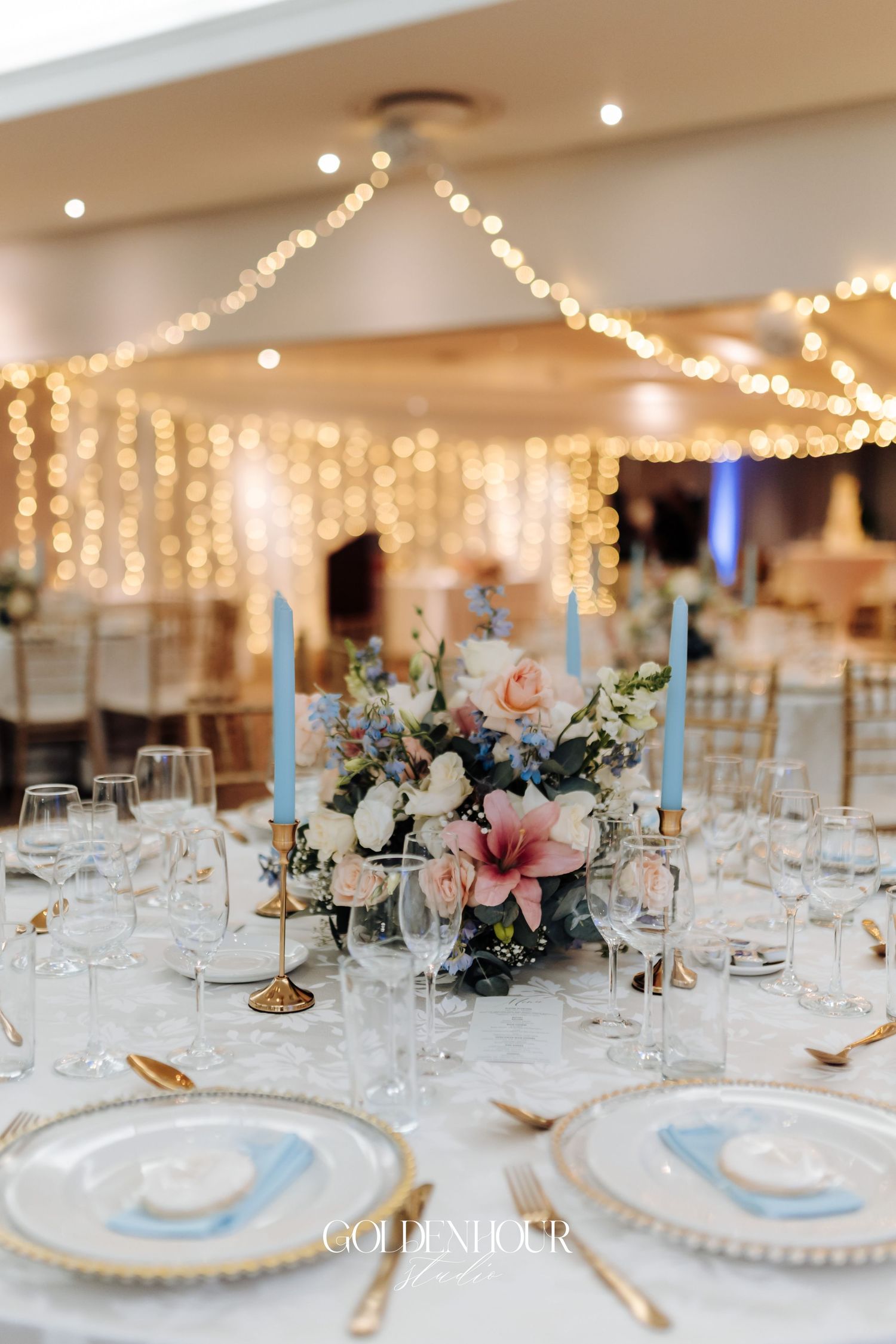 Wedding reception table setting with string lights and blue and pink floral centerpiece.