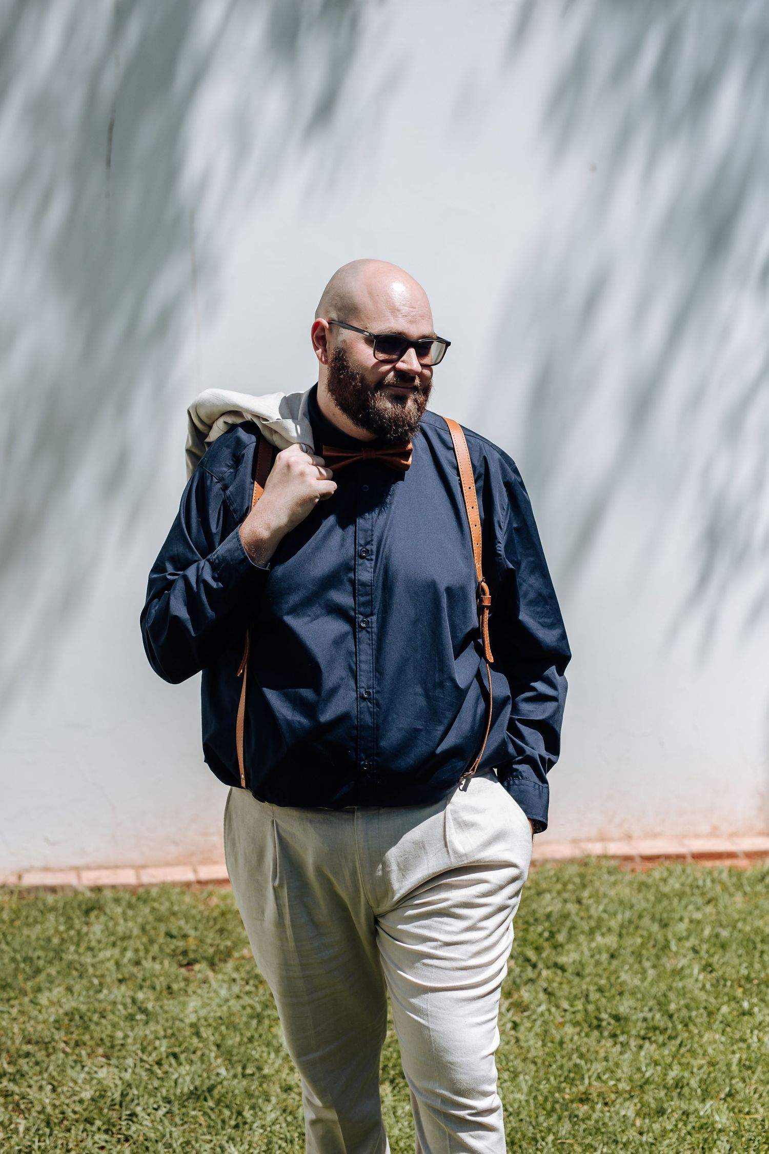 A person wearing sunglasses and a navy blue shirt with white pants walks on grass against a textured wall background.