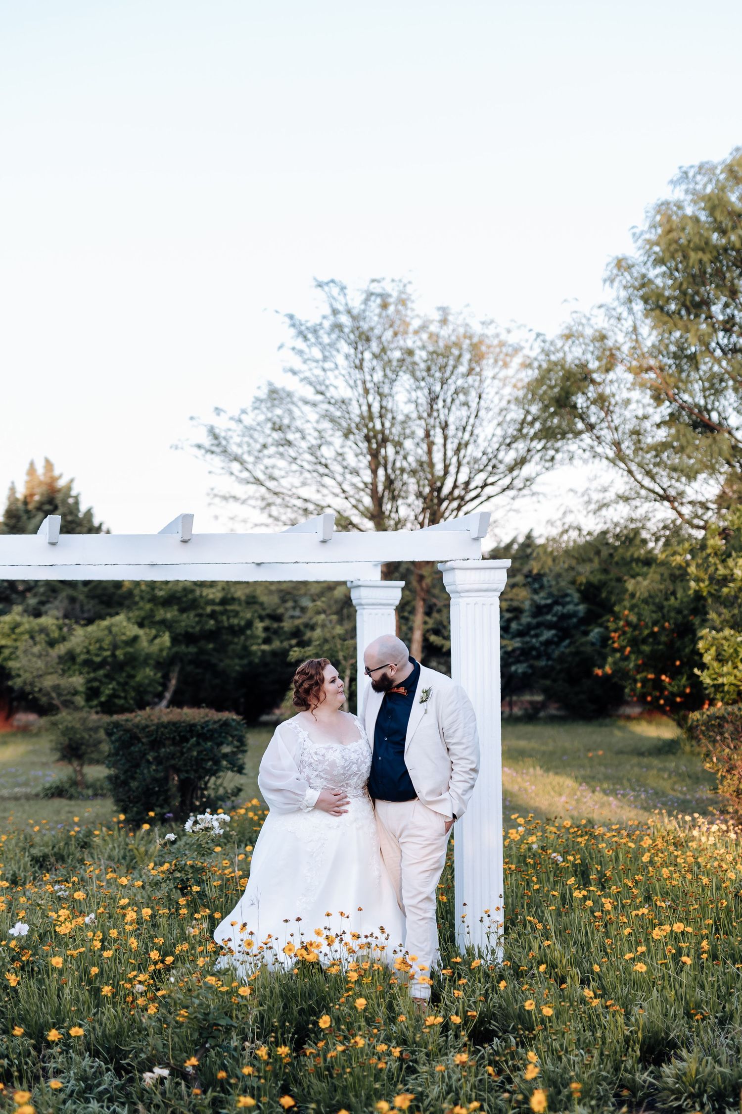 A couple in wedding attire sits together on a white bench surrounded by wildflowers in a meadow.