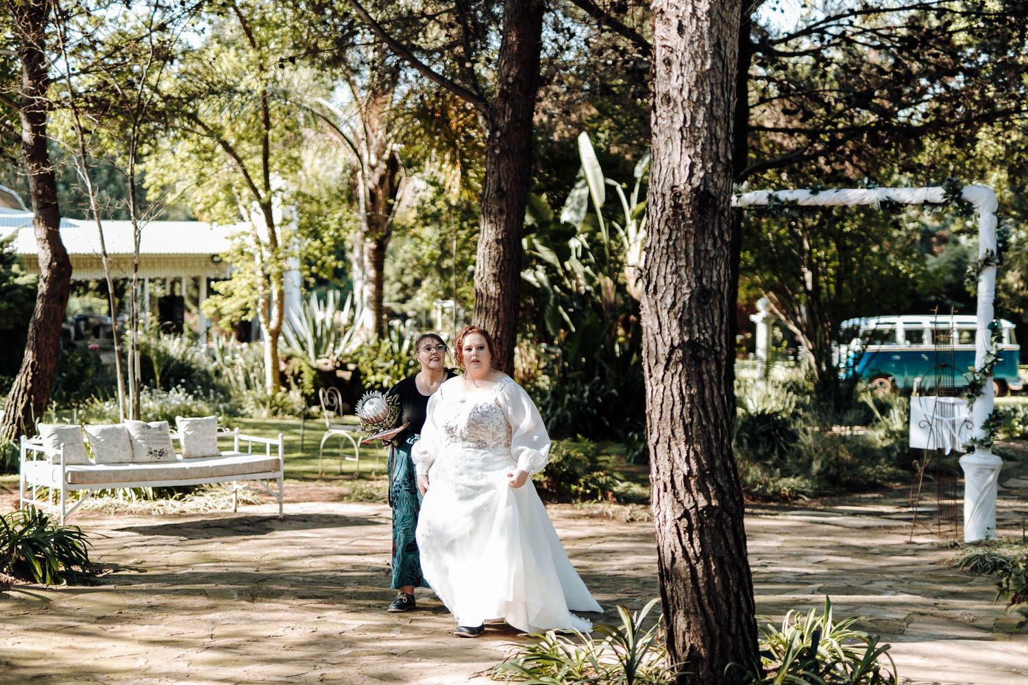 A bride in a flowing white dress runs through a sunlit garden path lined with tall trees and greenery.