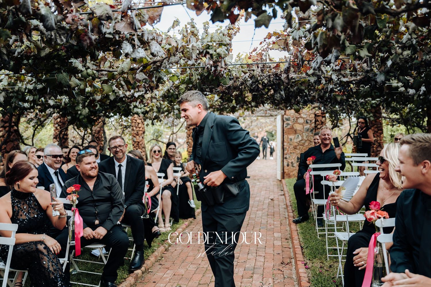 Outdoor wedding ceremony with guests seated on white chairs along a brick pathway surrounded by trees and greenery.