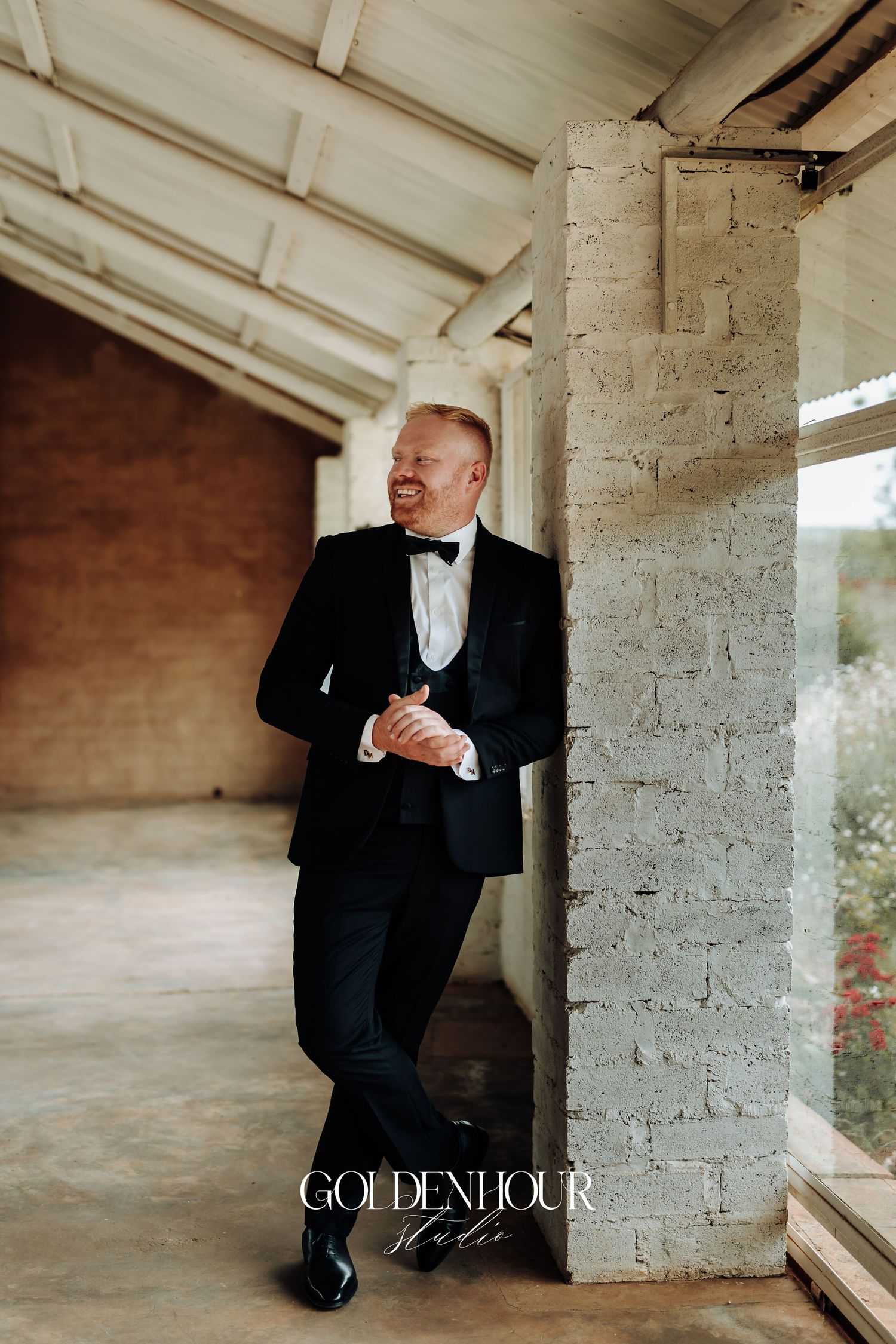 A person in a classic black tuxedo and bowtie leans against a rustic concrete pillar in an elegant wedding photo.
