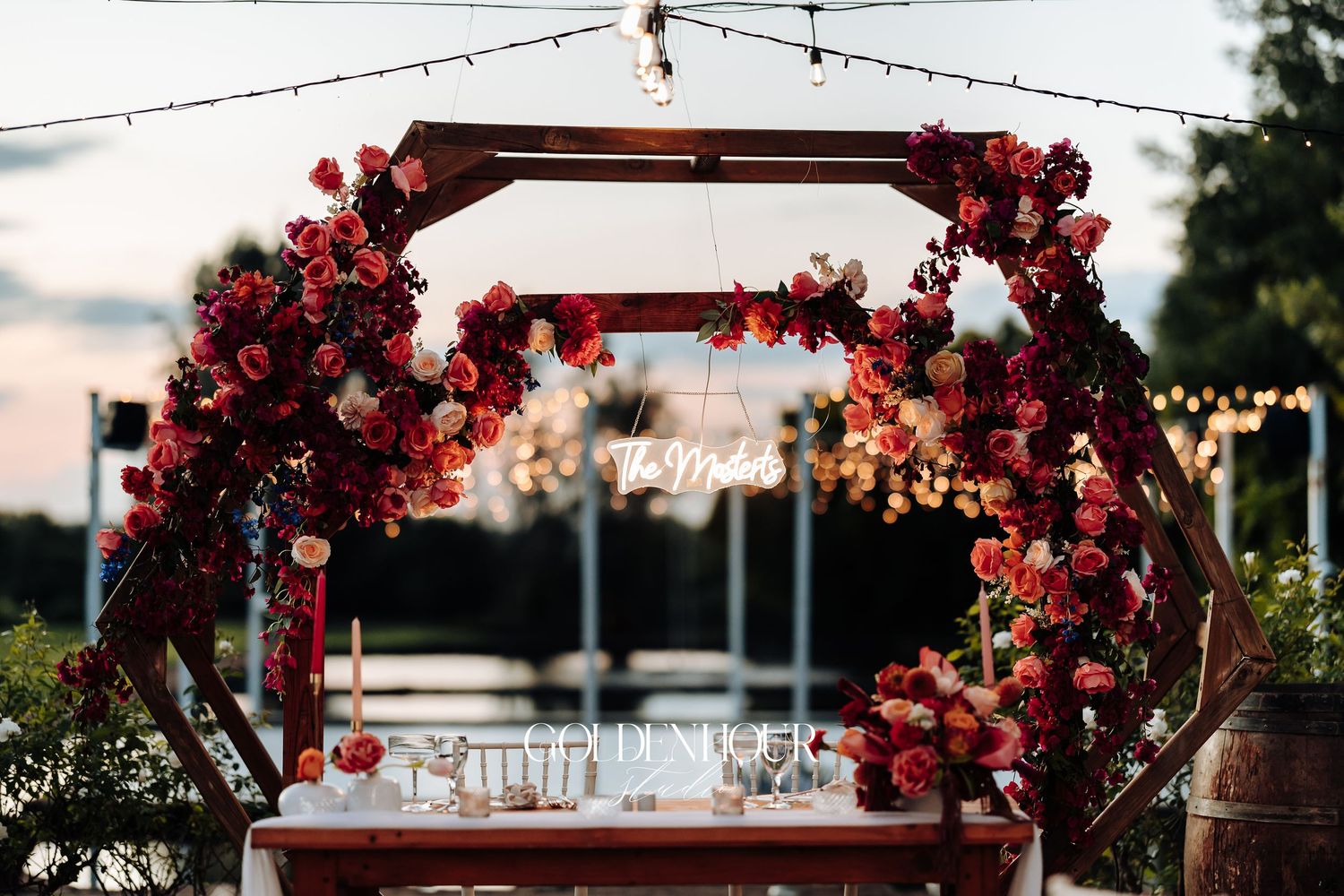 A wooden wedding arch decorated with red roses and floral arrangements against a sunset sky with string lights overhead.