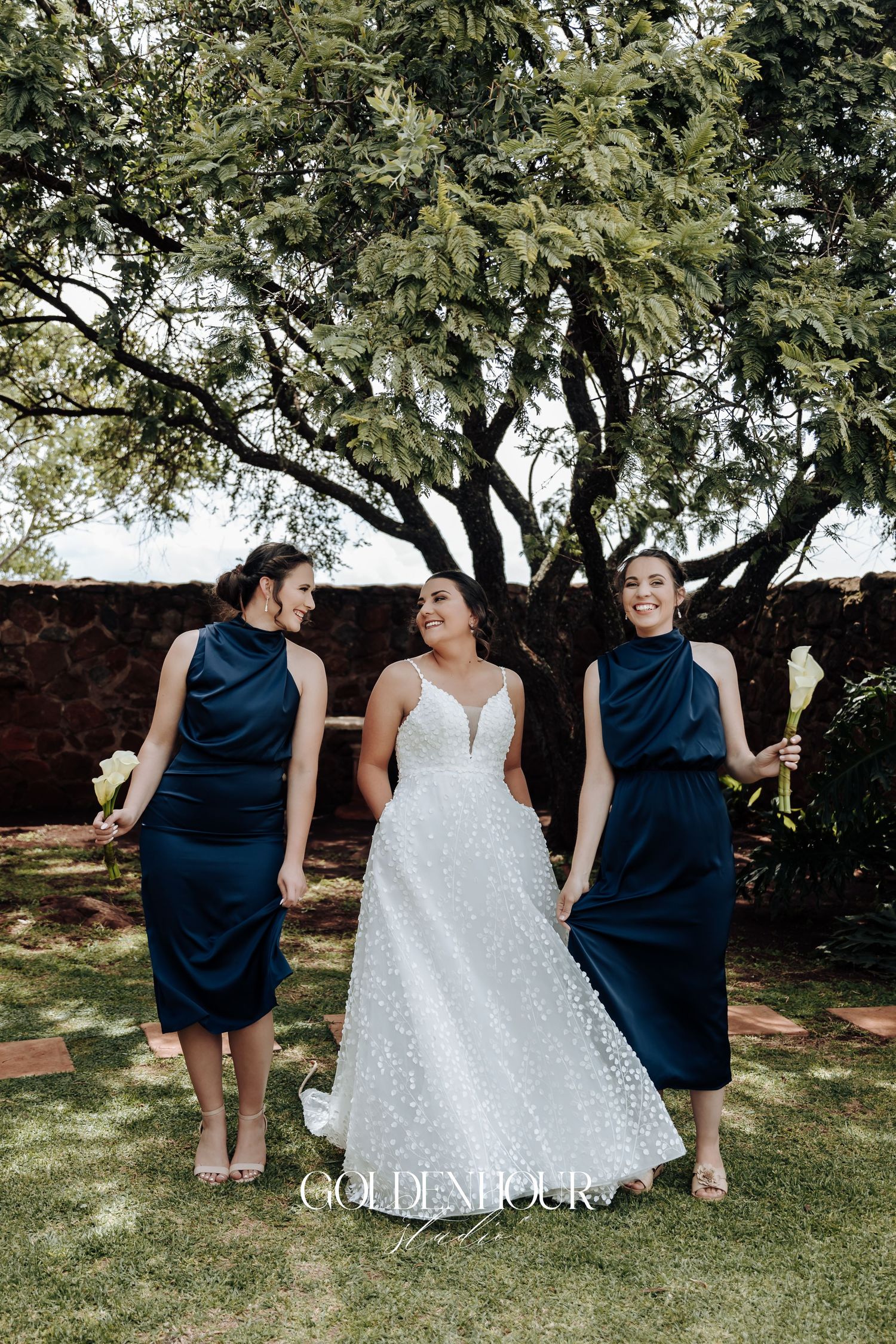 Wedding party members in navy and white dresses pose playfully on grass under trees.