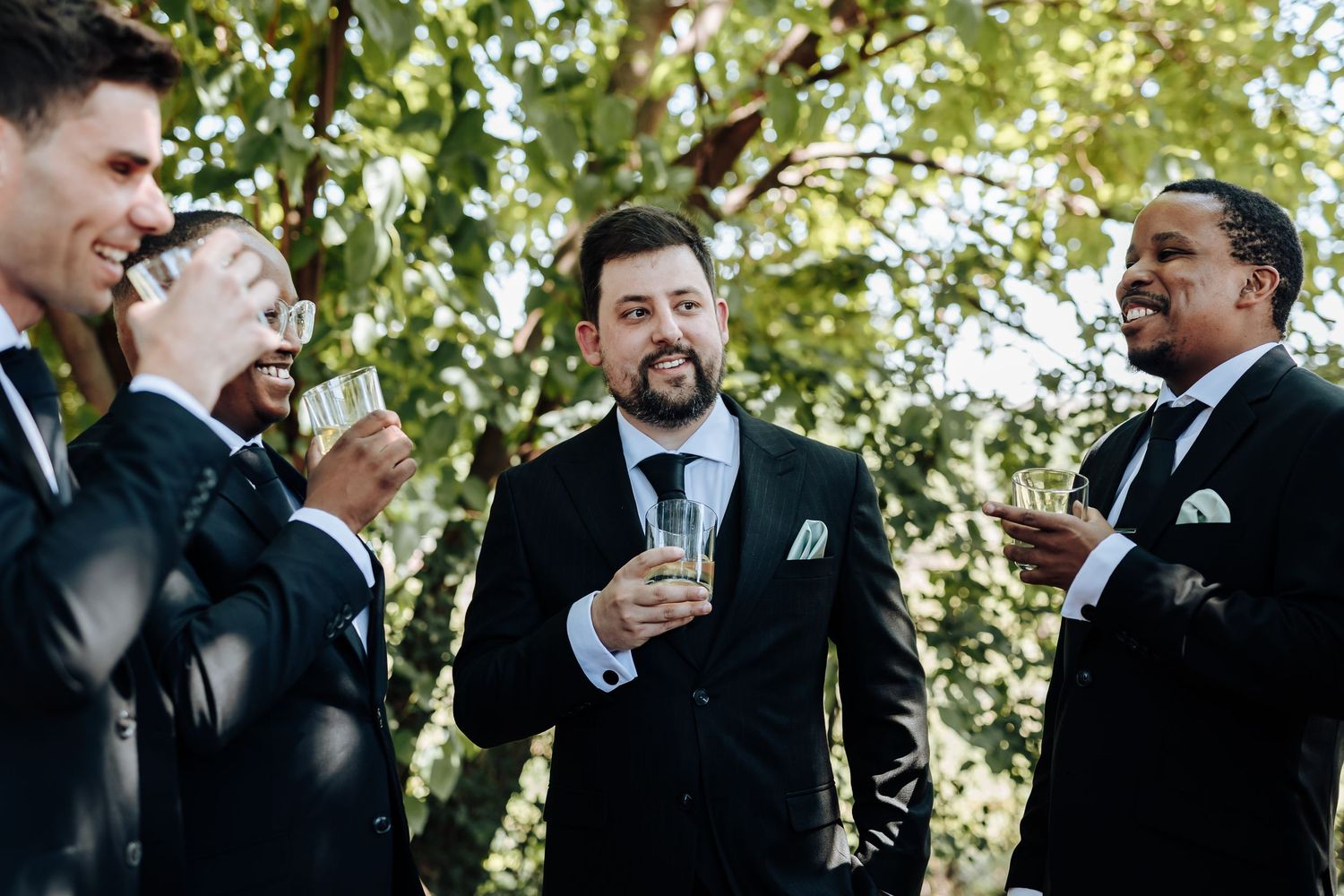 Wedding party members in formal attire gather for candid moments between trees in natural outdoor setting.
