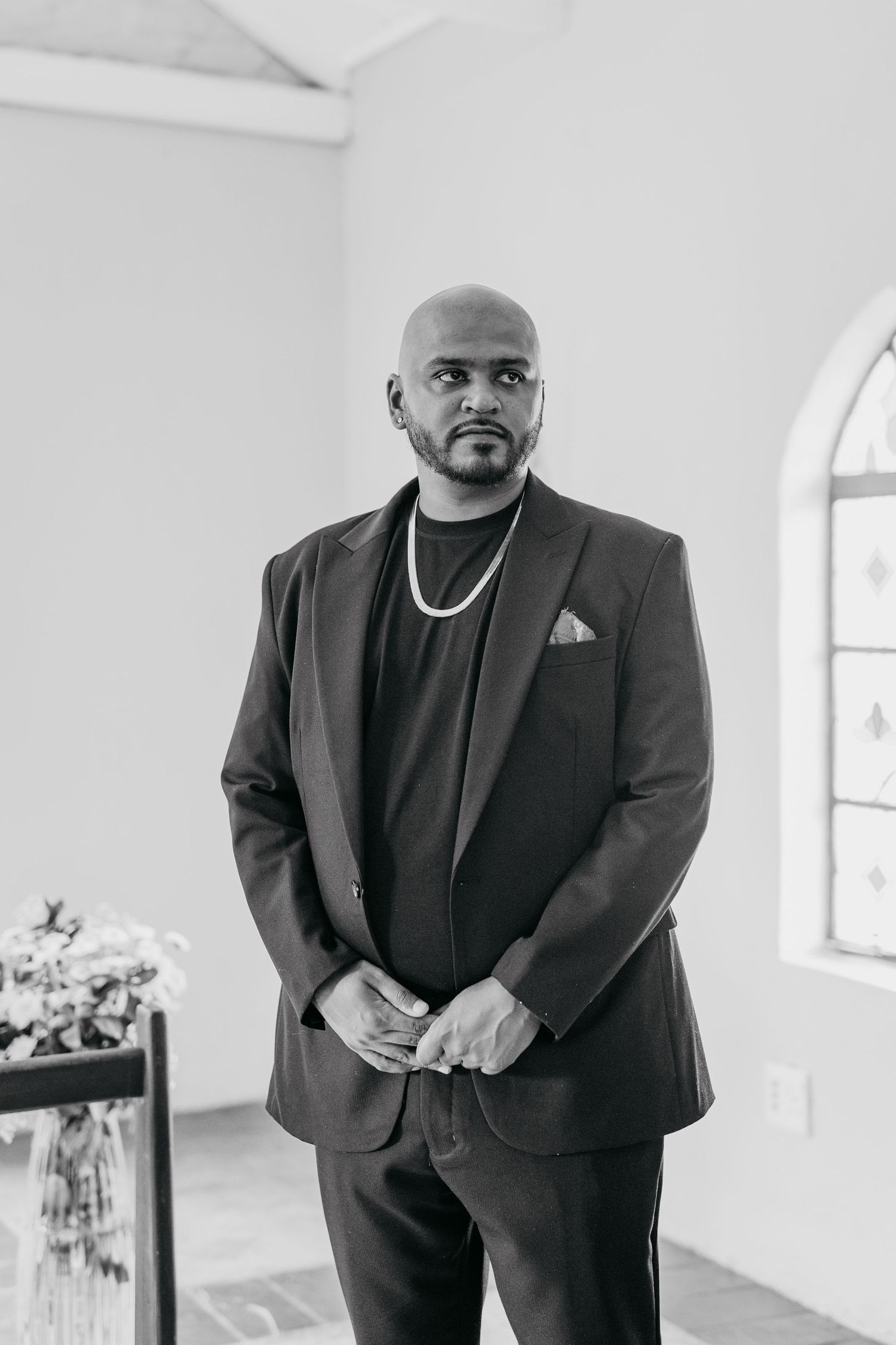 A formal portrait of a groom wearing a black suit with silver chain necklace in a bright white chapel.