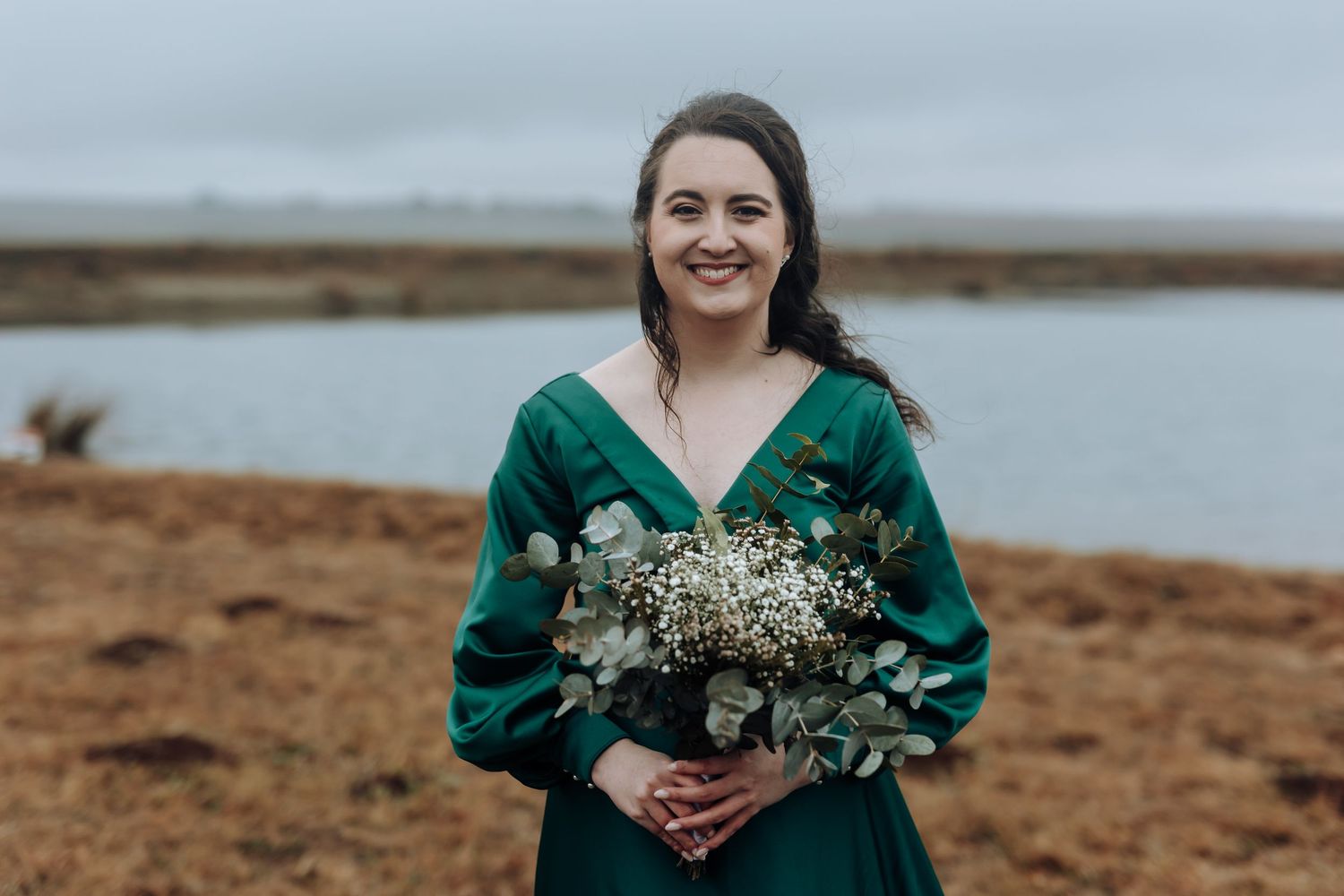 Bridesmaid in emerald dress holds white and green bouquet by waterfront.