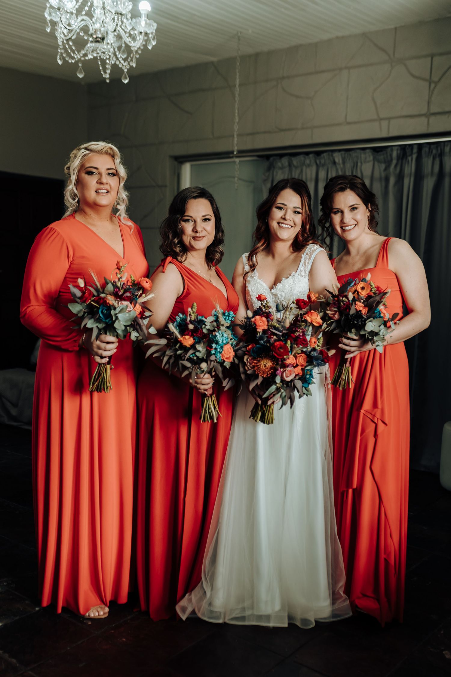 Bridal party poses under crystal chandelier in coral dresses holding vibrant bouquets with turquoise accents.