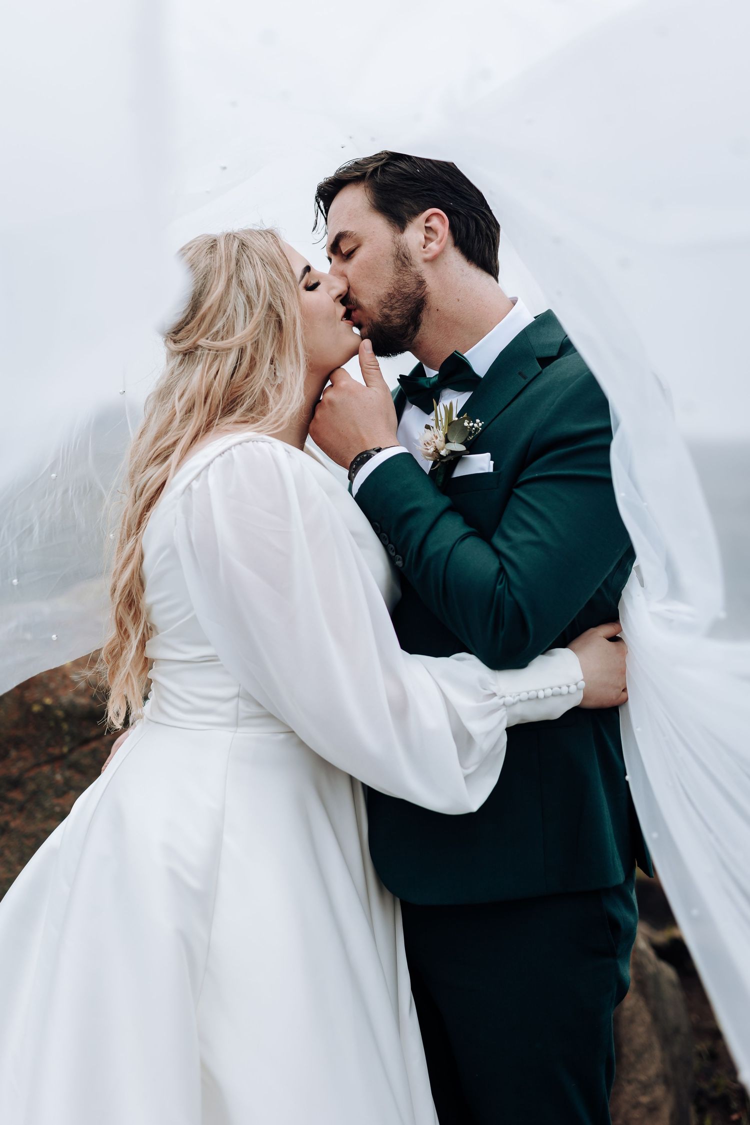 Intimate wedding kiss captured in elegant portrait with dramatic veil and emerald green suit.