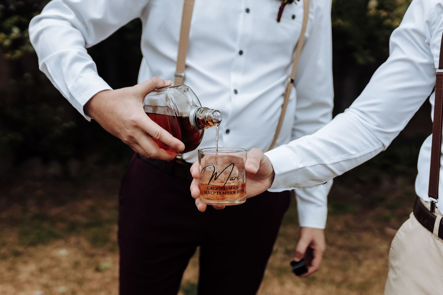 Wedding party members in suspenders pour whiskey into crystal glasses.