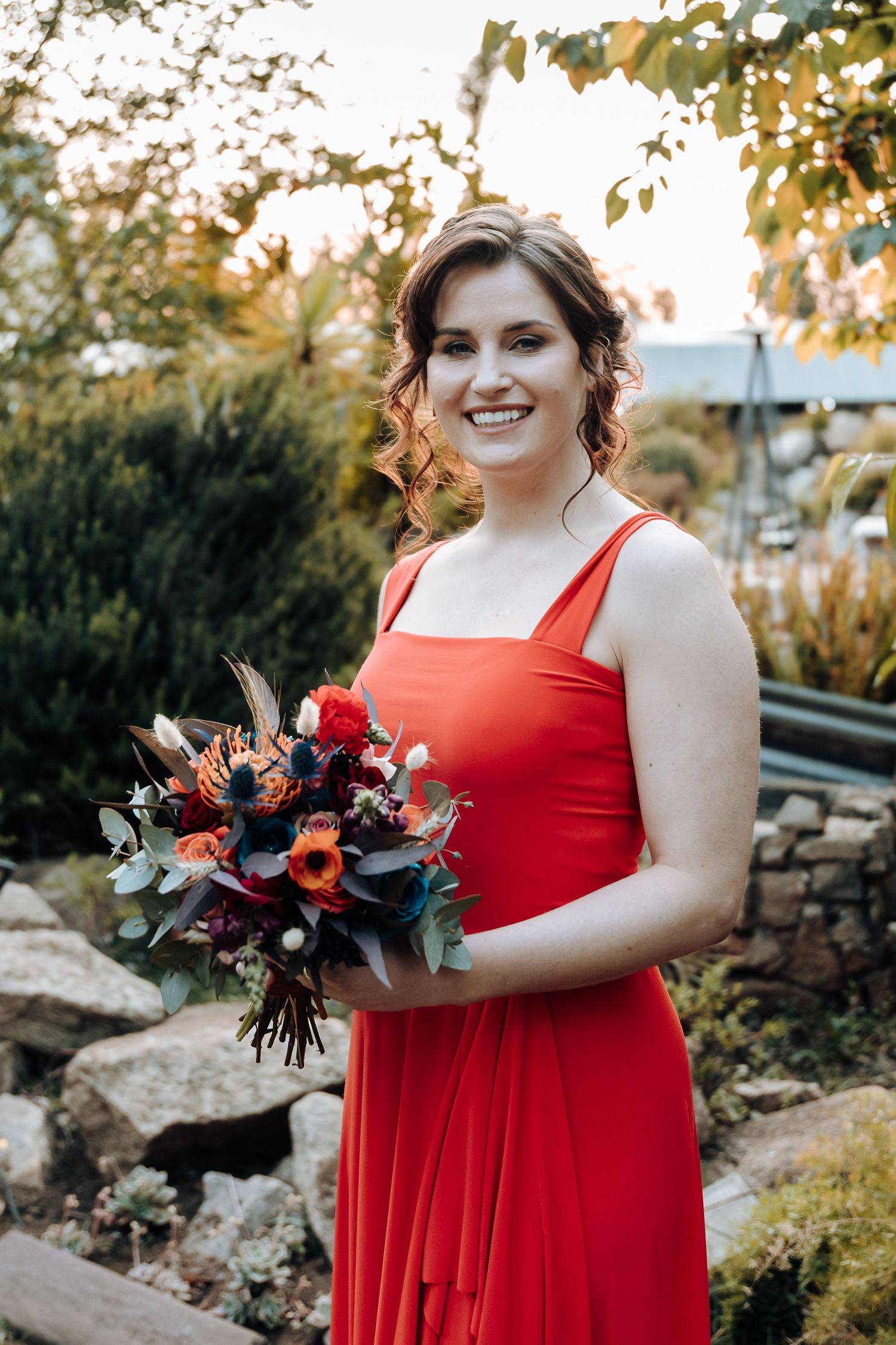 Bridesmaid in red dress holds colorful bouquet while posing in garden setting.