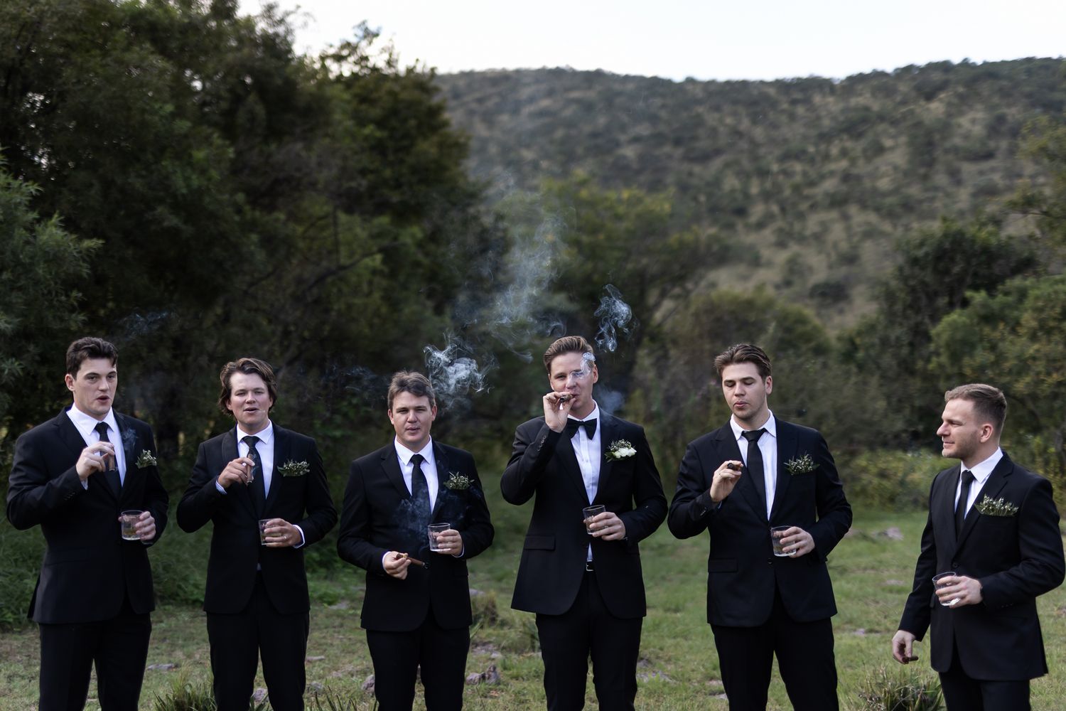 Groomsmen in formal attire smoking cigars at dusk with mountains in the background.