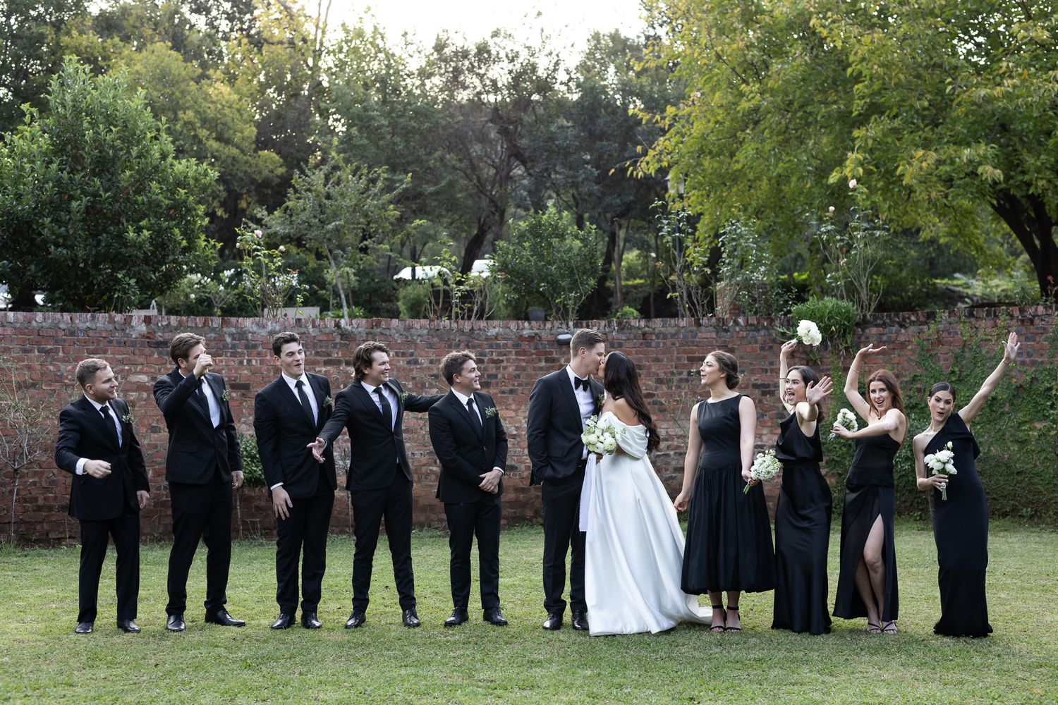 A wedding party in black attire poses together on a grassy lawn with a stone wall backdrop.
