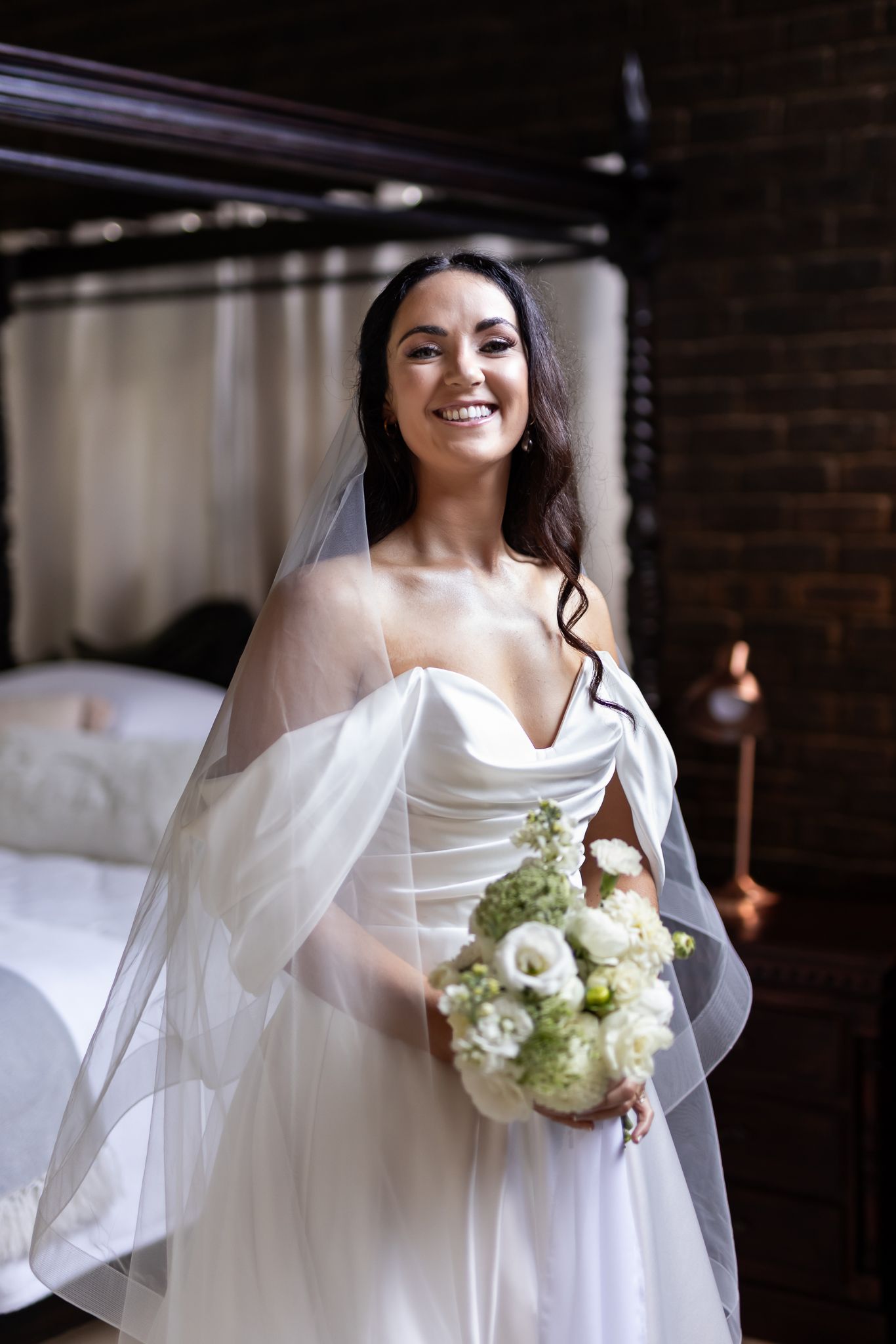 Bride in off-shoulder dress holding white bouquet poses next to canopy bed.