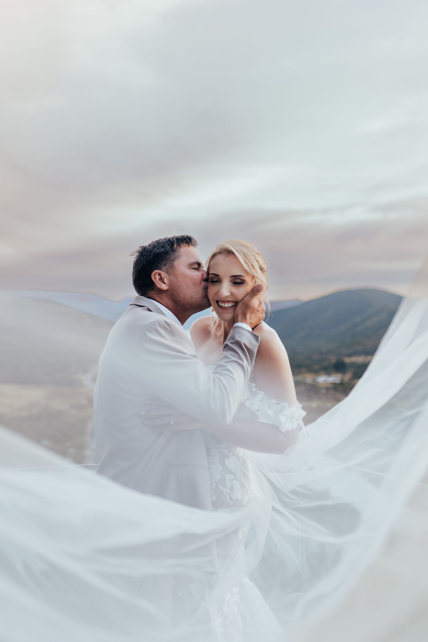 A romantic wedding portrait with flowing white veil against a mountain landscape at sunset.