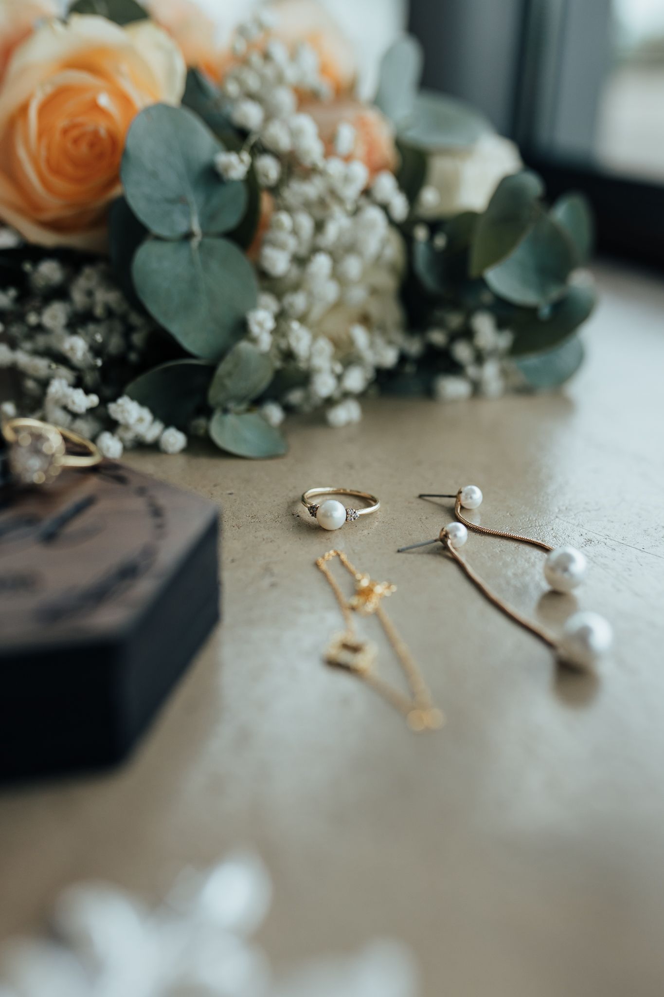 Wedding jewelry and floral arrangement with peach roses and eucalyptus on wooden surface.