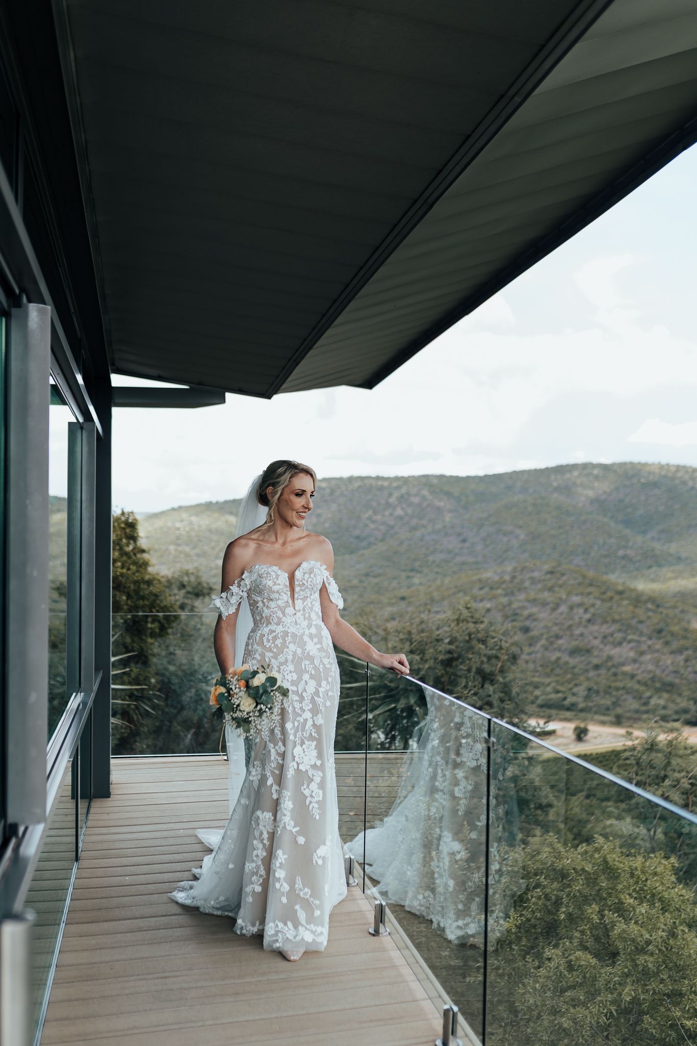 Bride in off-shoulder lace gown stands on modern balcony overlooking mountain landscape.