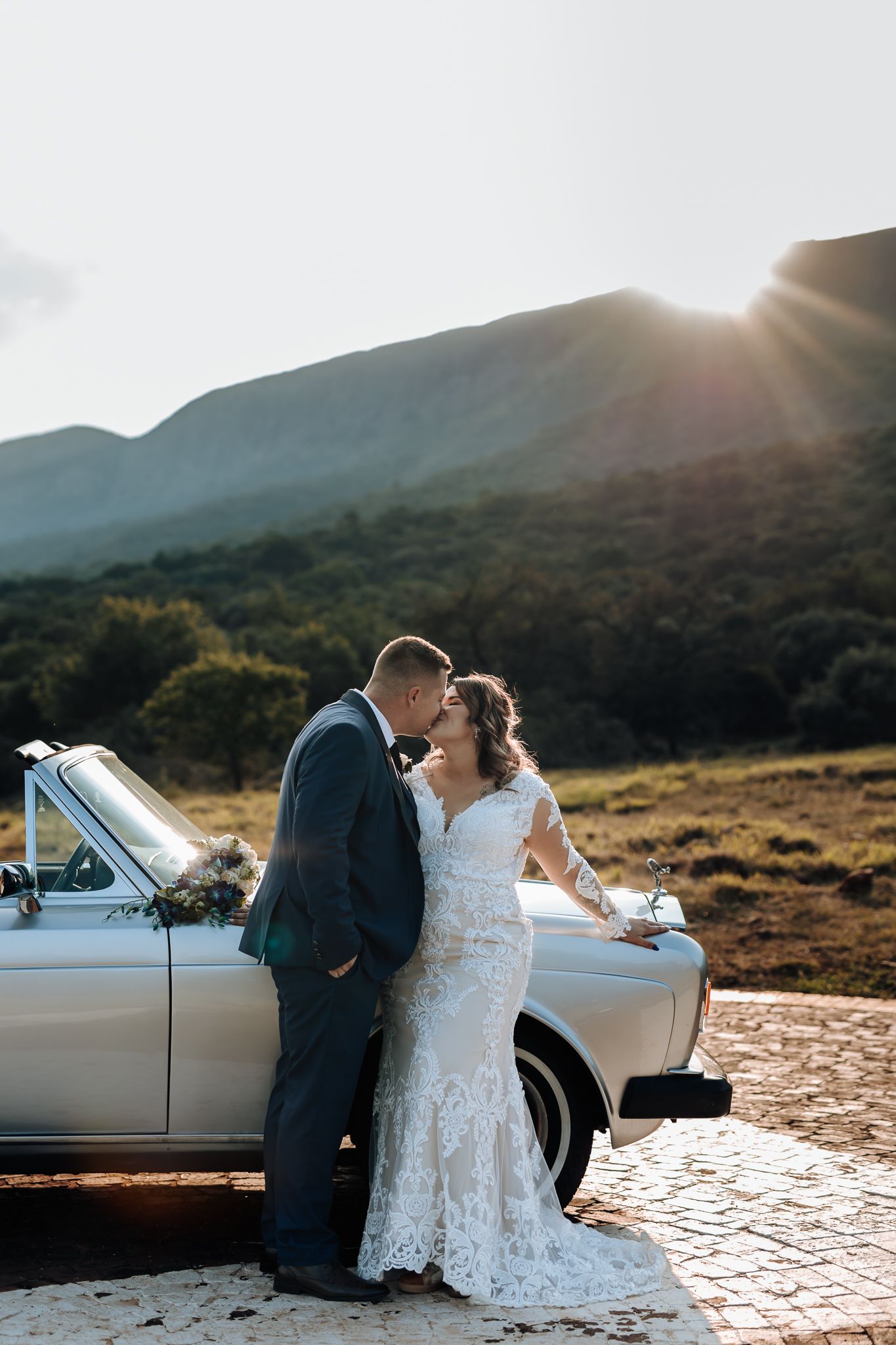 Romantic wedding photo with vintage car against mountain landscape at golden hour.