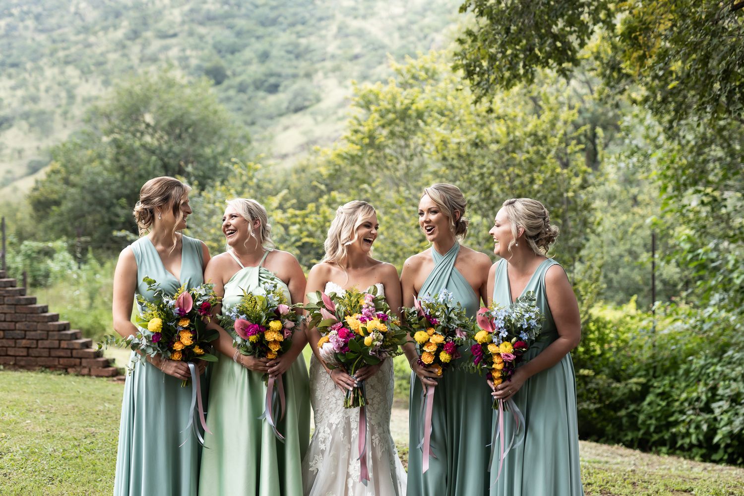 Wedding party in flowing mint dresses poses with bright floral bouquets on a hillside.