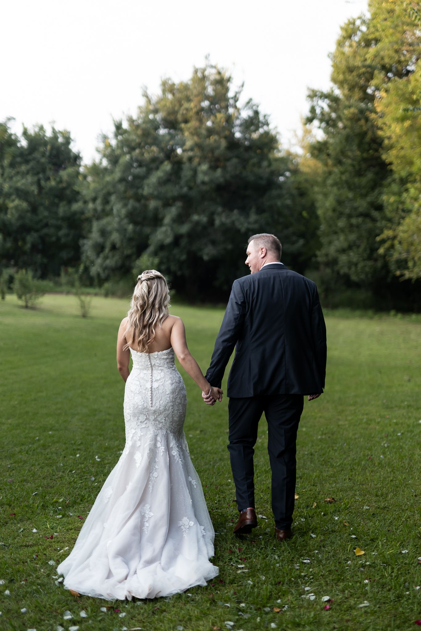 A newlywed couple walks hand in hand through a lush garden setting in wedding attire.