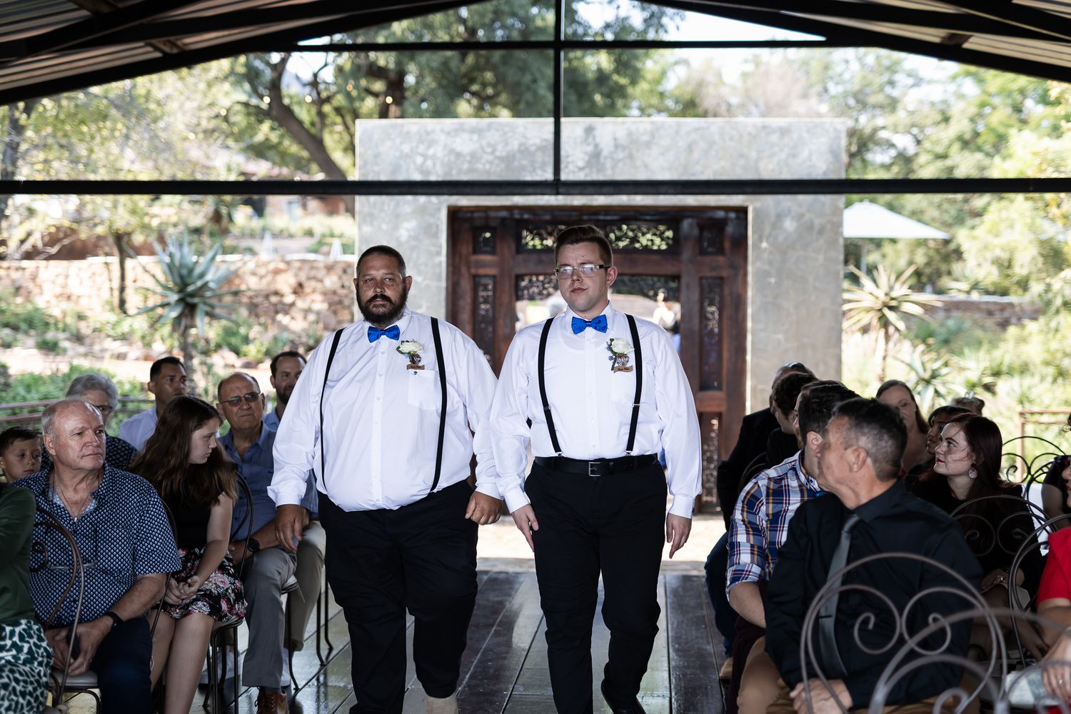 Two groomsmen in black suits with suspenders walk down the aisle during a wedding ceremony.