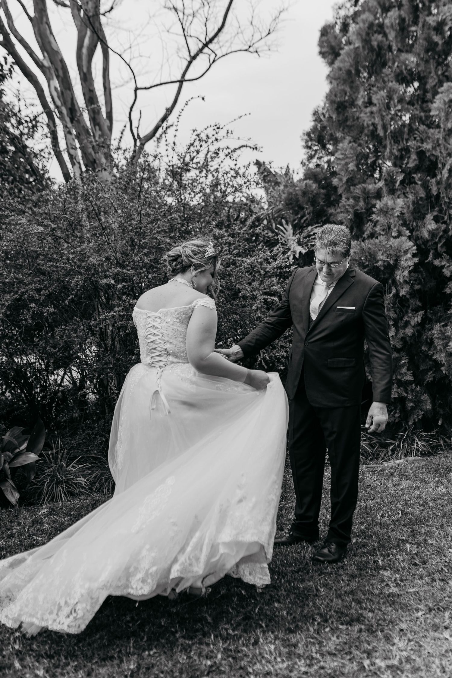 A black and white photograph shows a couple in wedding attire standing together on grass near trees and bushes.