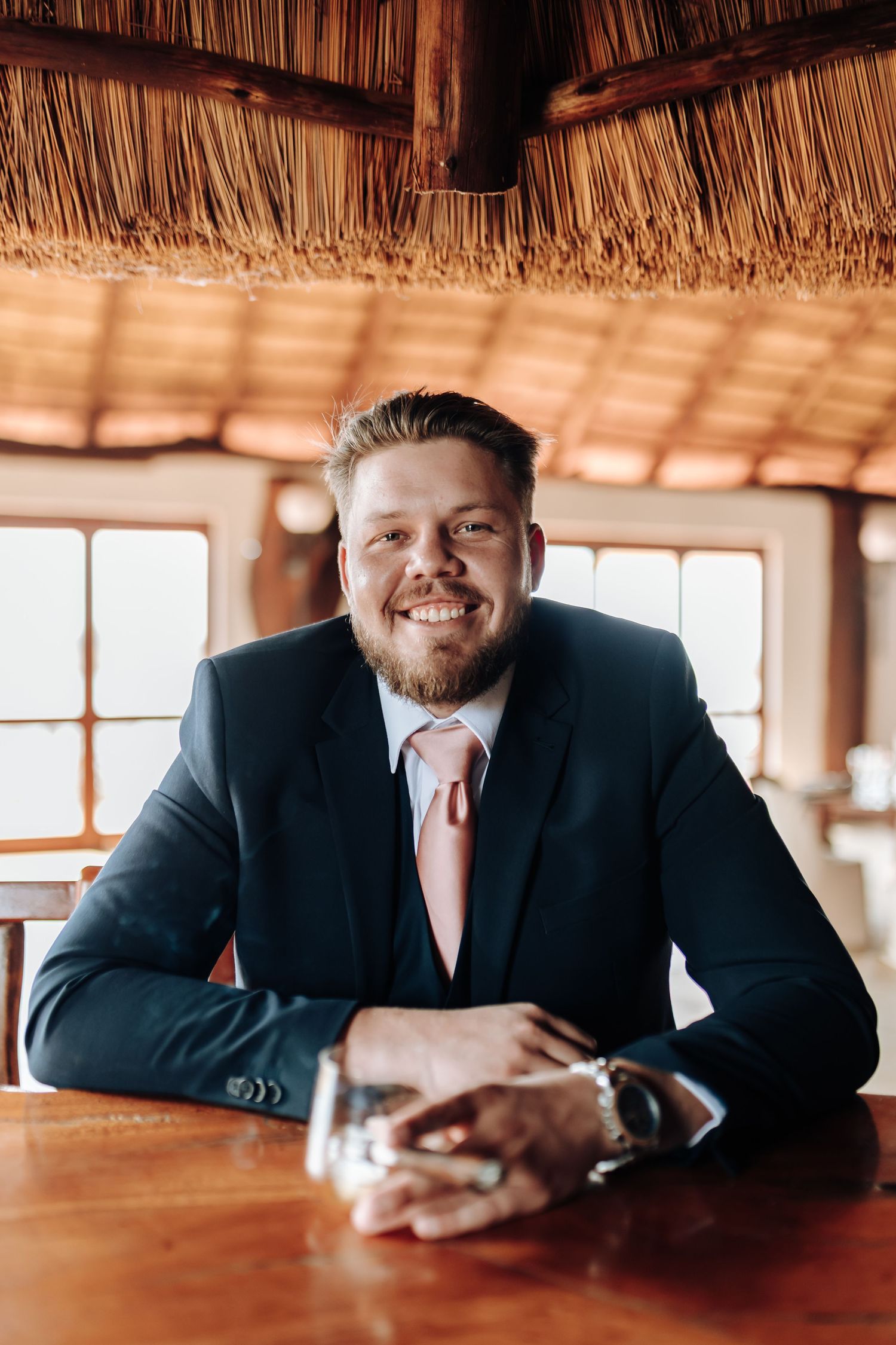 Professional in a navy suit smiles while relaxing on wooden table in rustic tiki-style venue with thatched roof decor.