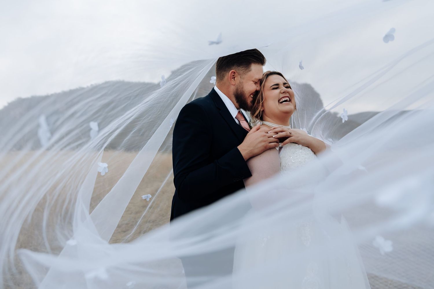 A bride and groom share an intimate moment under a flowing white veil against a soft background.