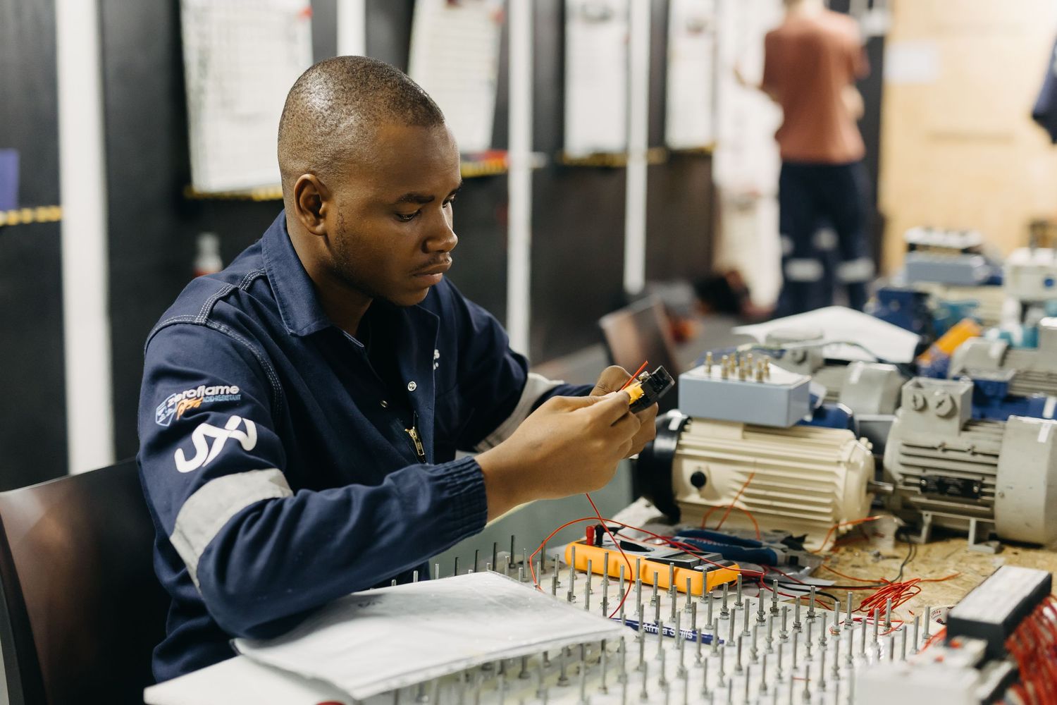 Engineers in navy uniforms inspect electronic components at a manufacturing workstation.
