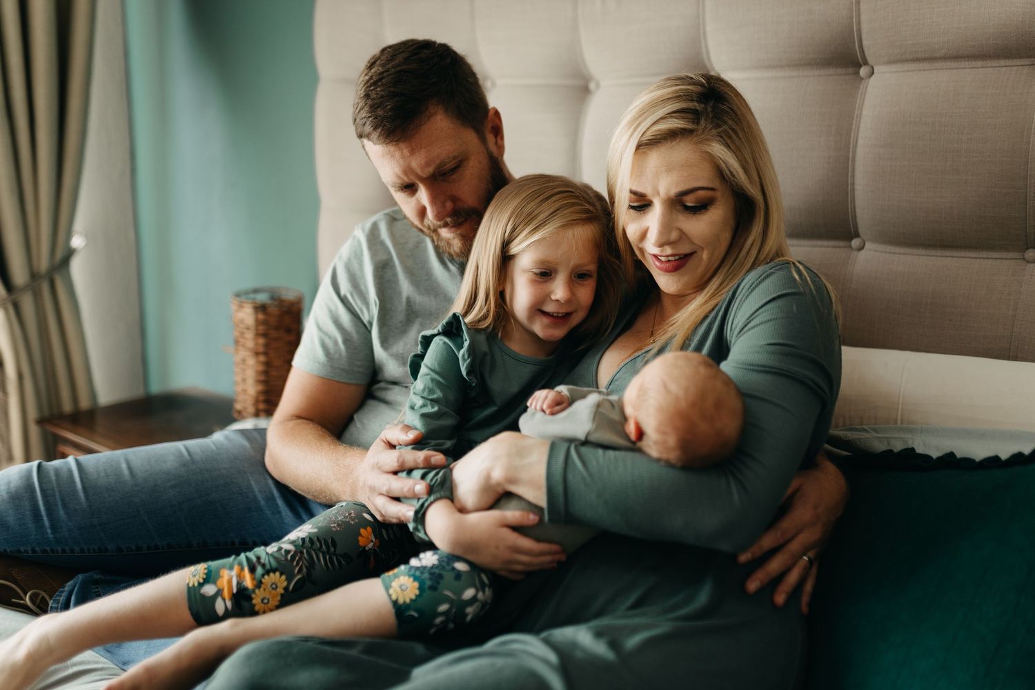 A family relaxes together on a bed, sharing a tender moment with their children in casual indoor setting.