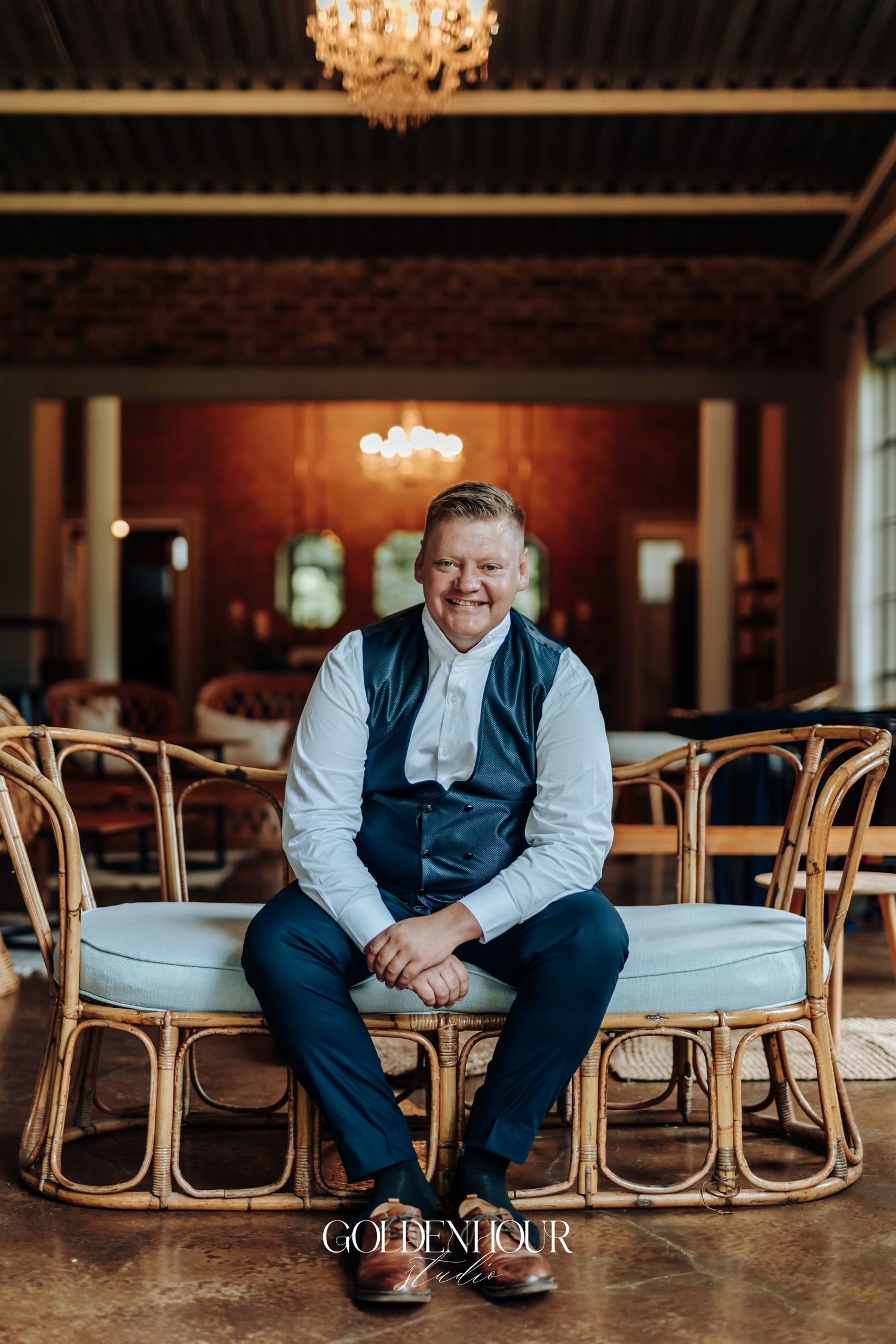 Smiling groom in navy vest and blue trousers seated on rattan furniture in a warmly lit wedding venue.
