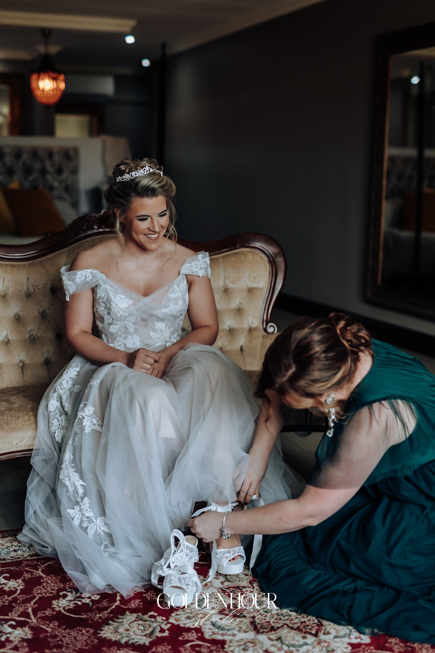 Smiling bride in lace gown seated on vintage sofa while bridesmaid helps with wedding shoes.