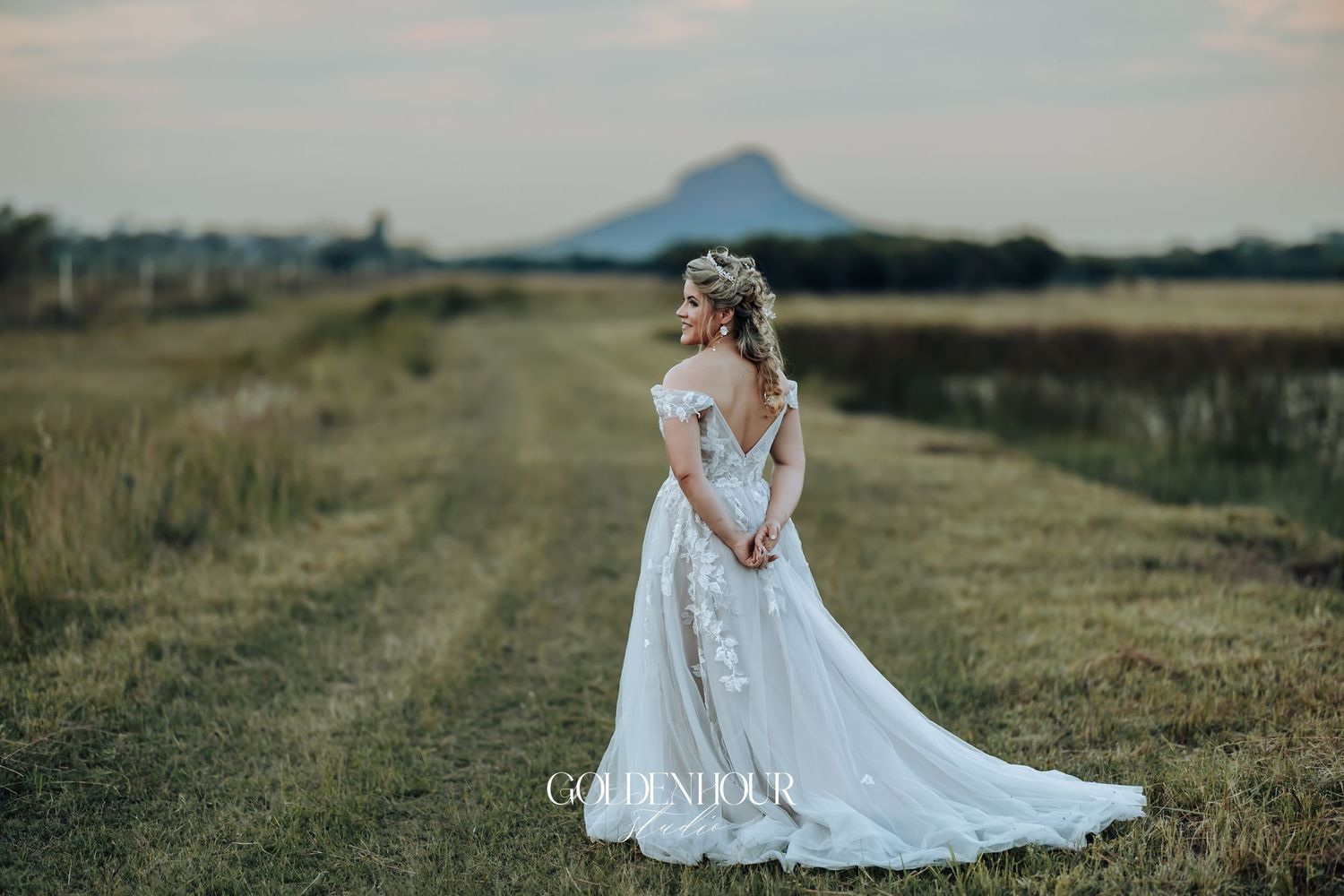 Bride in lace wedding gown stands on rural dirt path with mountain backdrop at dusk.
