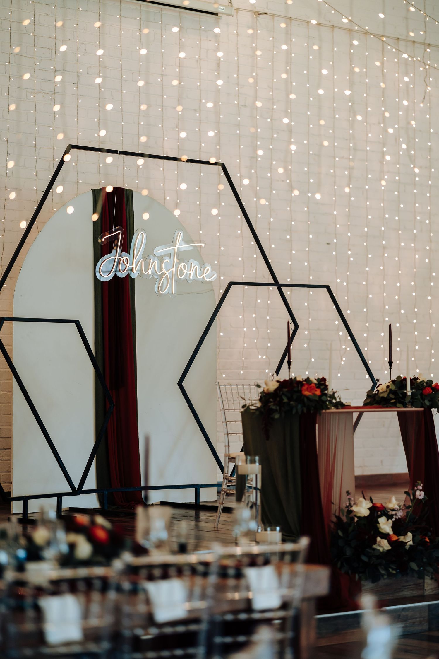 Elegant wedding reception backdrop with hexagonal frames, fairy lights, neon Johnstone sign, and floral arrangements on draped table.