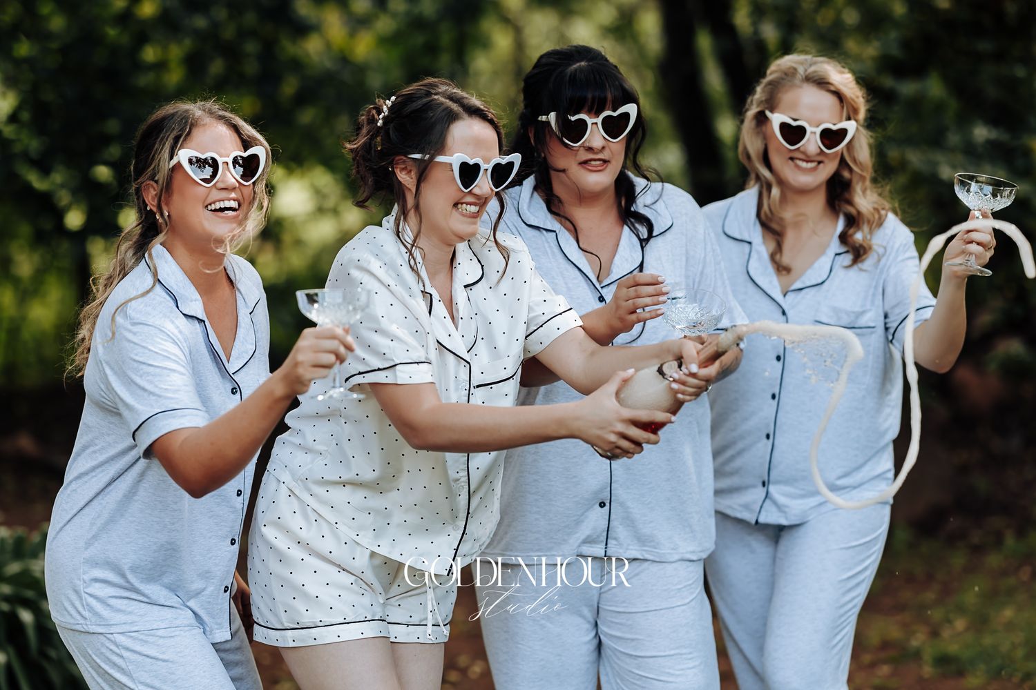 Four bridesmaids in blue pajamas wearing heart sunglasses popping champagne outdoors.