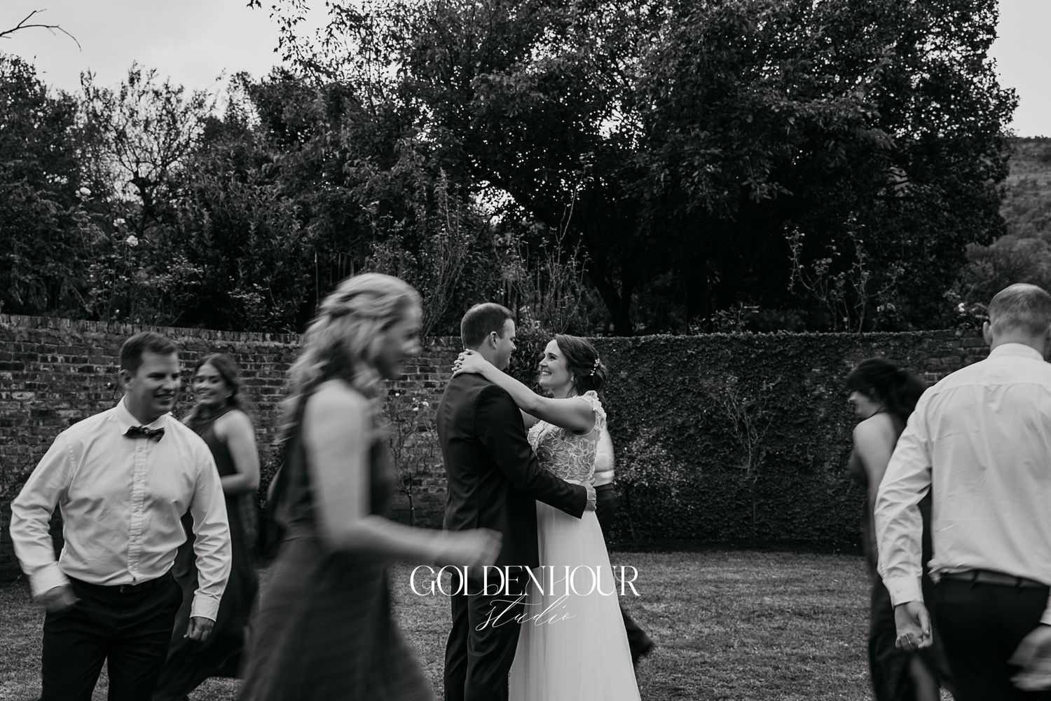 Bride and groom share a romantic moment outdoors with wedding guests nearby.