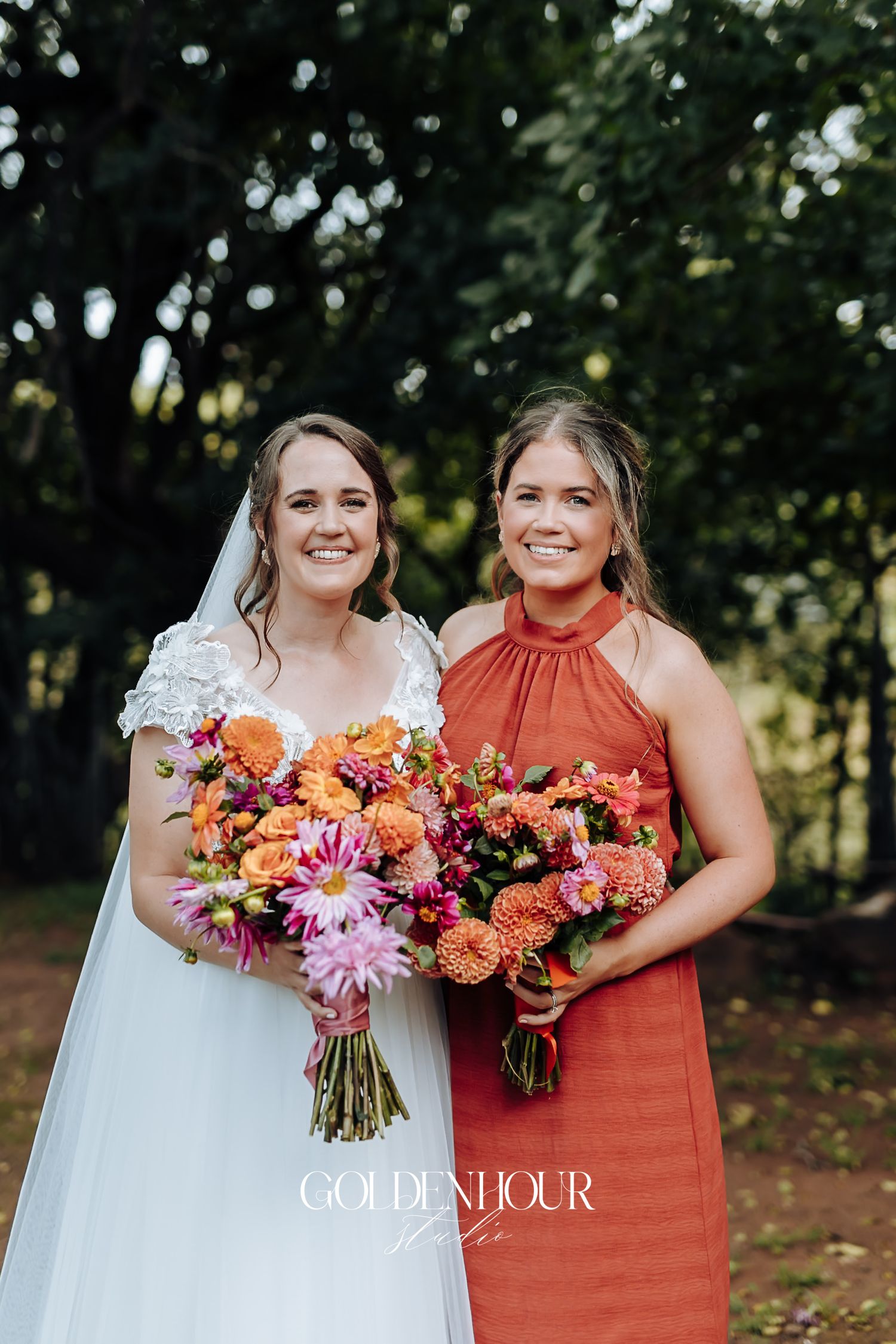 Bride and bridesmaid in rust dress holding matching orange and pink floral bouquets outdoors.